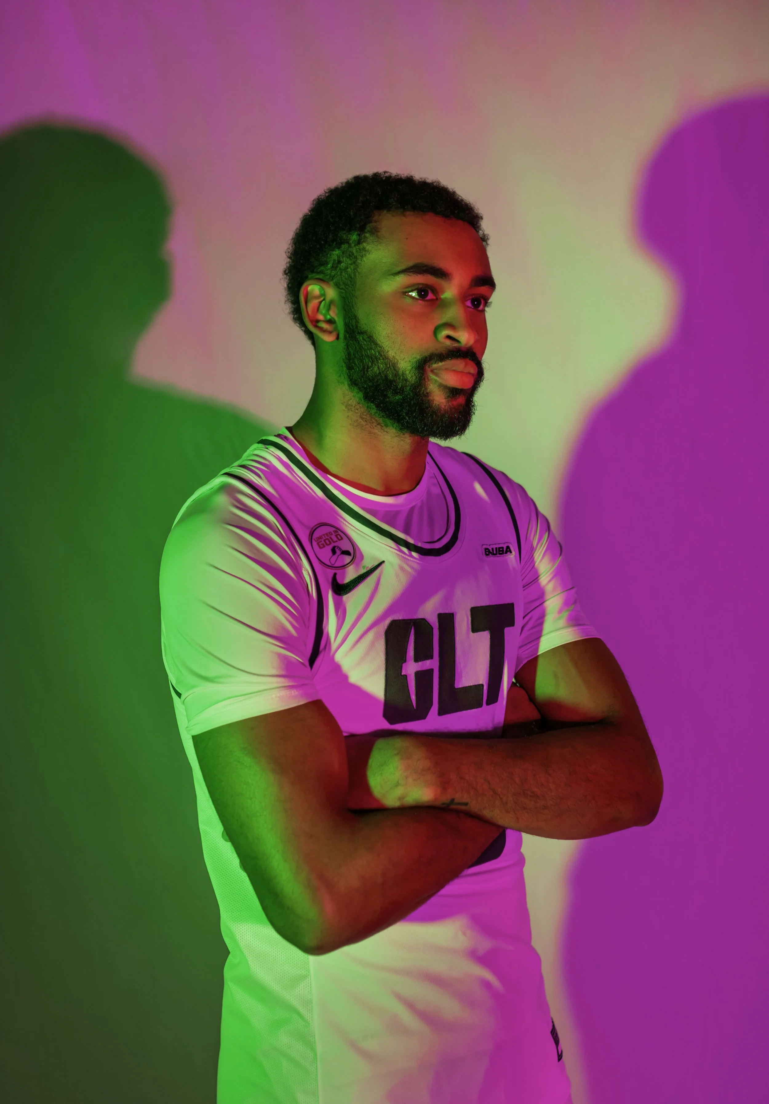 A man in a white sports jersey with black lettering, crossing arms, standing against a wall with colorful pink, green, and purple lighting creating shadows.