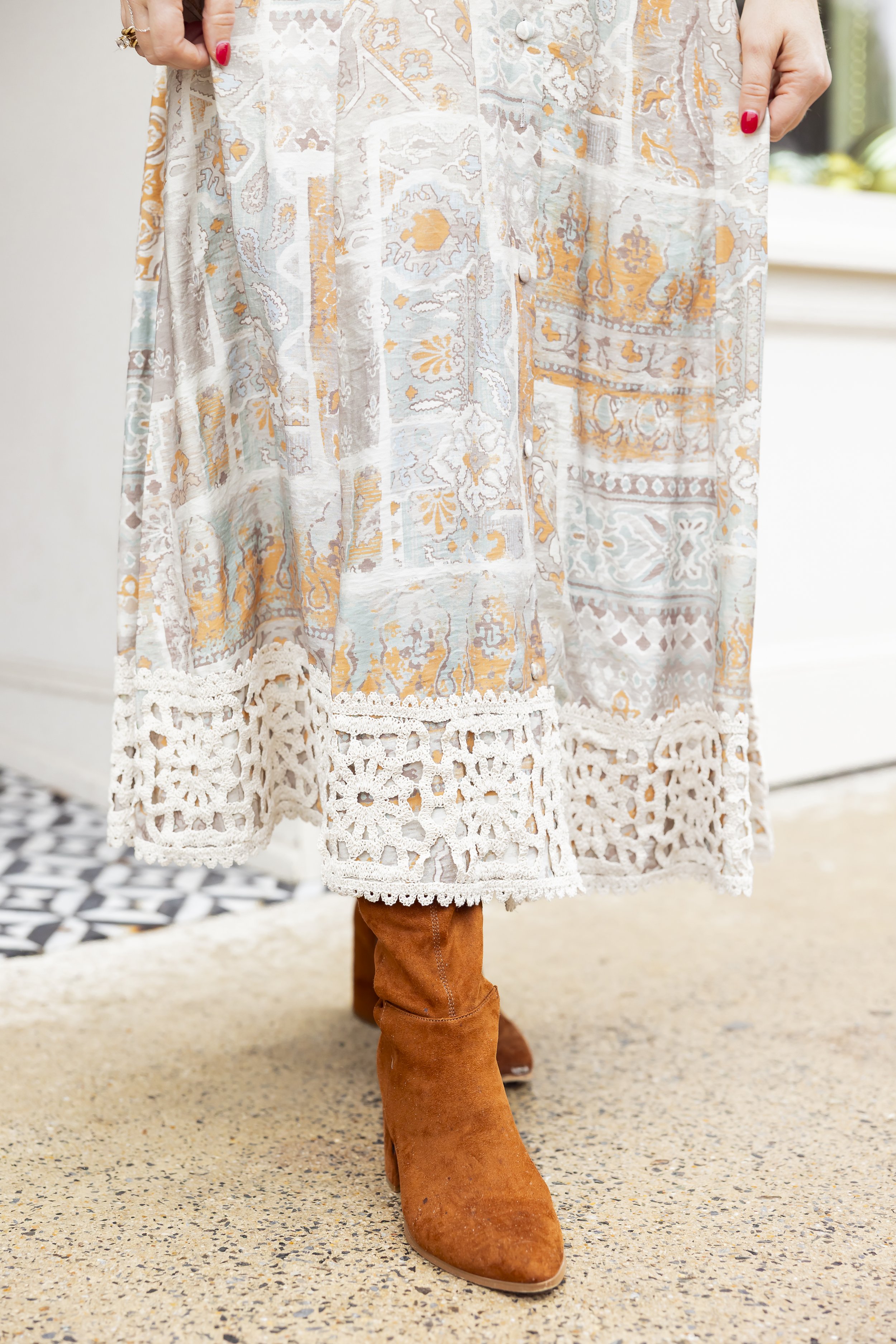 Close-up of a person wearing brown suede boots, a long patterned dress with crochet lace at the hem, and holding the dress with one hand. The background shows a room with a tiled floor and white walls.