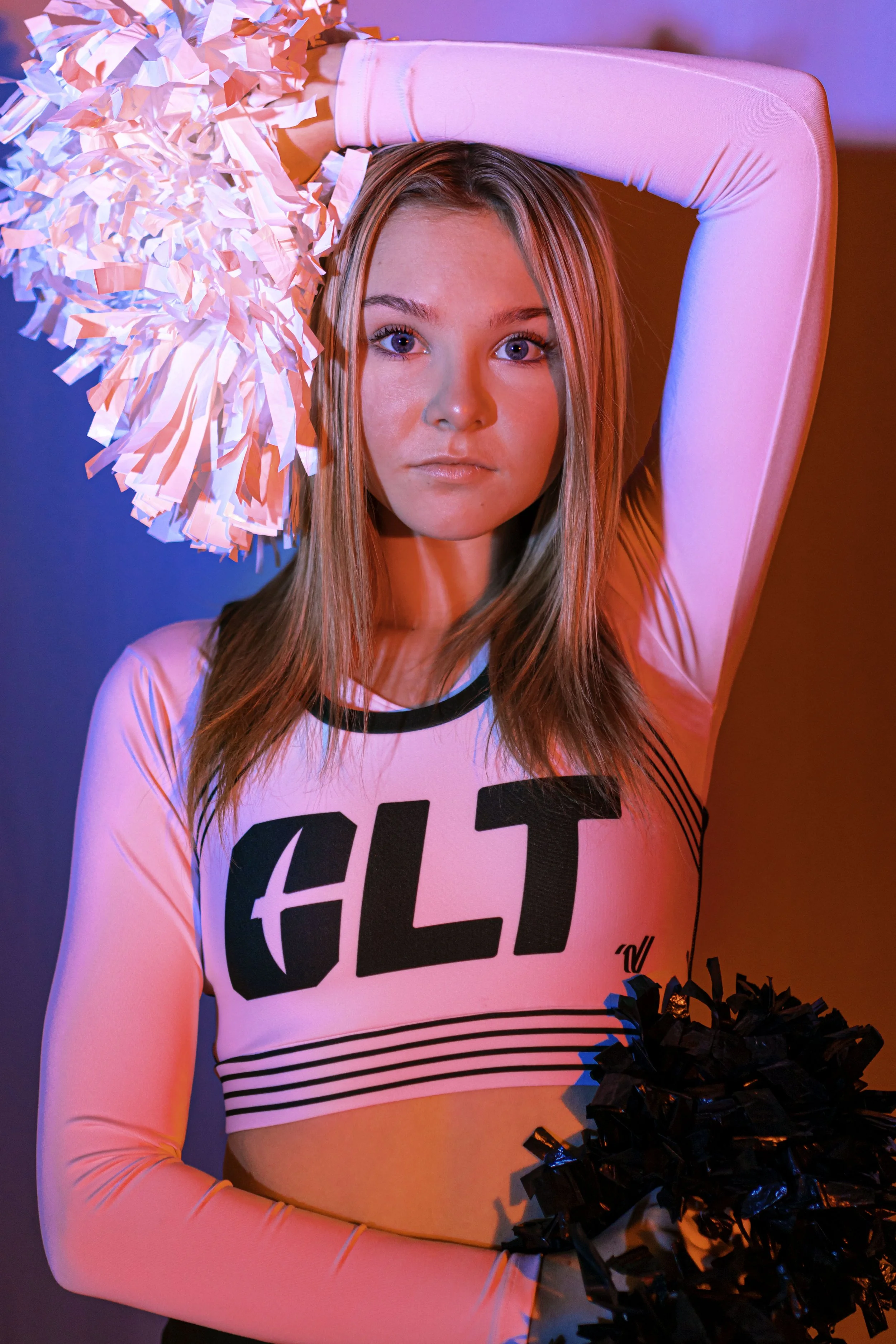 A cheerleader with long blonde hair in a white and black cheerleading uniform holds black pom-poms. She is standing in front of a background lit with pink and blue lights.