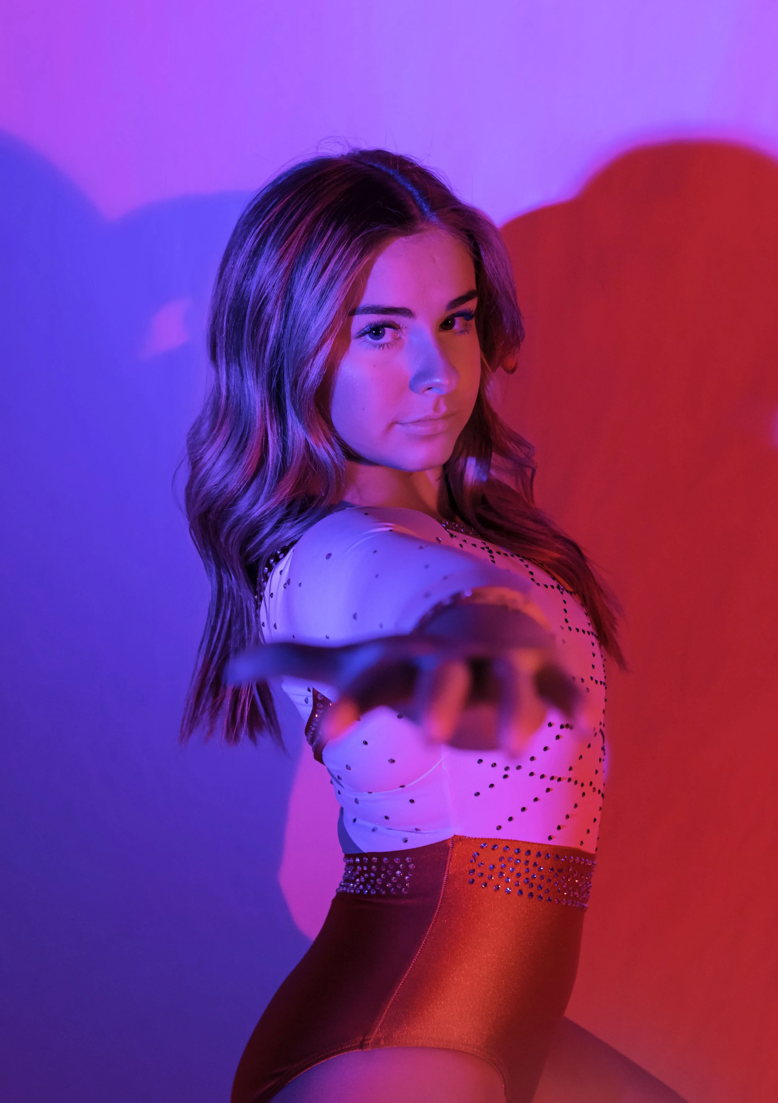 Young woman with wavy brown hair posing in colorful purple, blue, and red lighting, wearing a white and red outfit with black embellishments, reaching towards the camera.