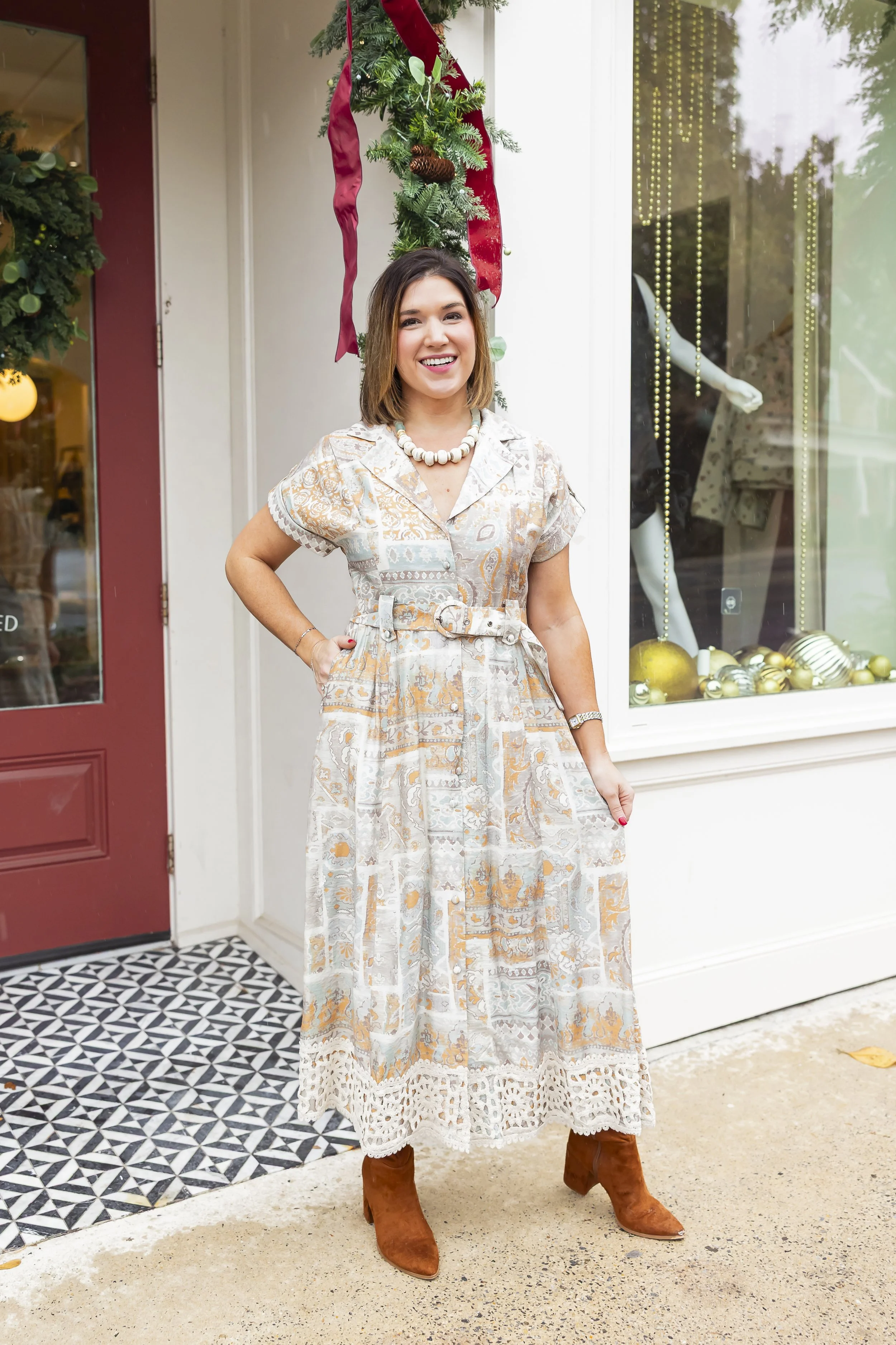 A woman standing outside a decorated storefront during Christmas, wearing a patterned dress, brown boots, and jewelry, smiling at the camera.