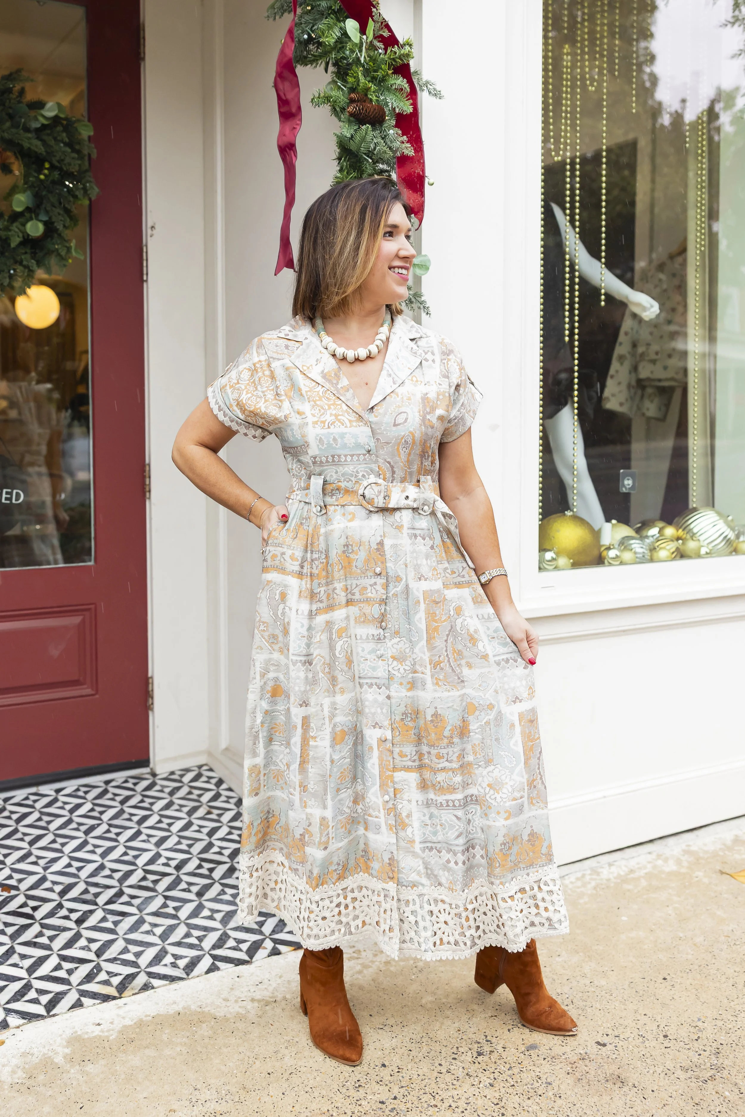 Woman wearing a long patterned dress and brown boots standing outside a store decorated with Christmas ornaments, with a Christmas wreath and greenery overhead, smiling and looking to the right.