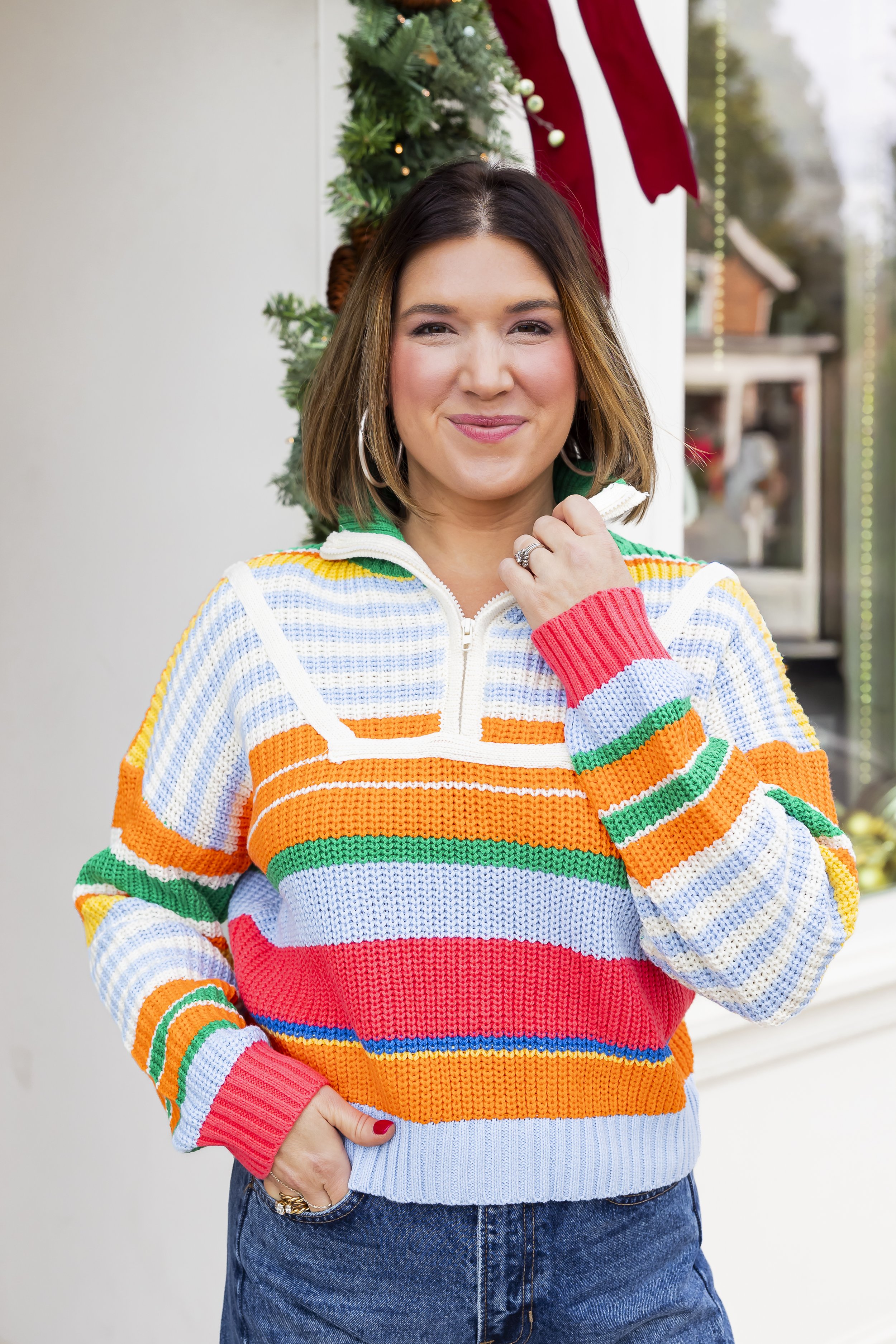 A woman with shoulder-length brown hair smiling, wearing a colorful, striped sweater and standing in front of a Christmas wreath with a red bow and other holiday decorations.