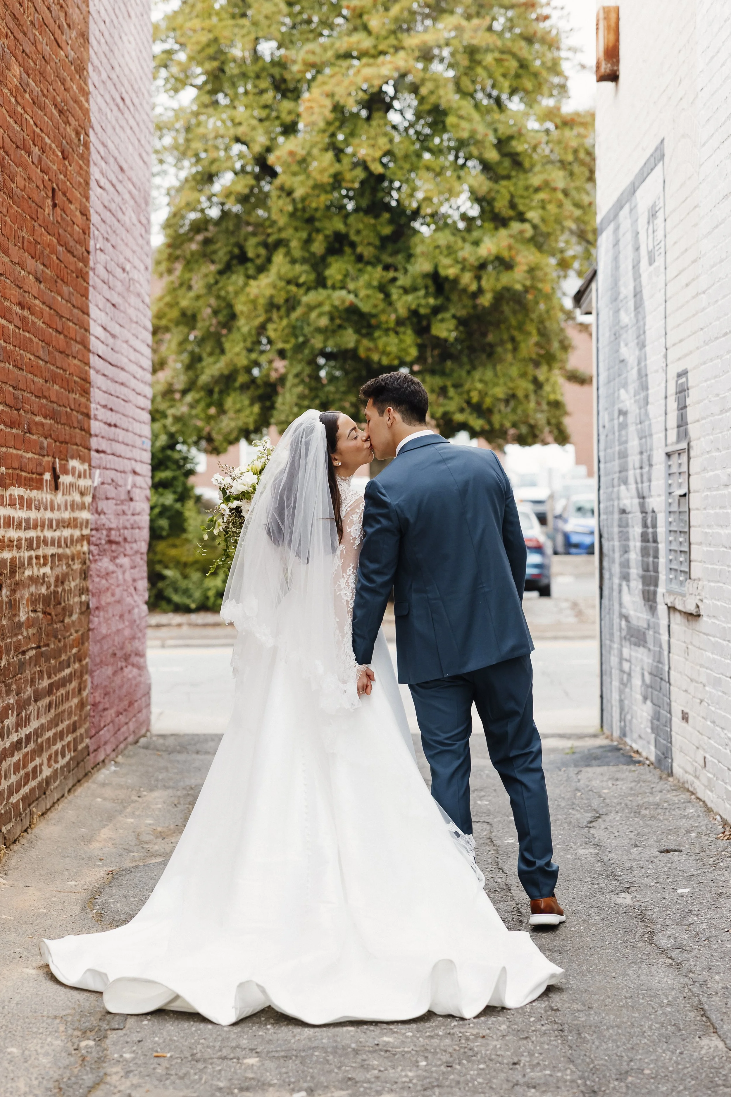 Bride and groom sharing a kiss in an alleyway with brick walls on either side, a large tree in the background, and parked cars visible beyond.