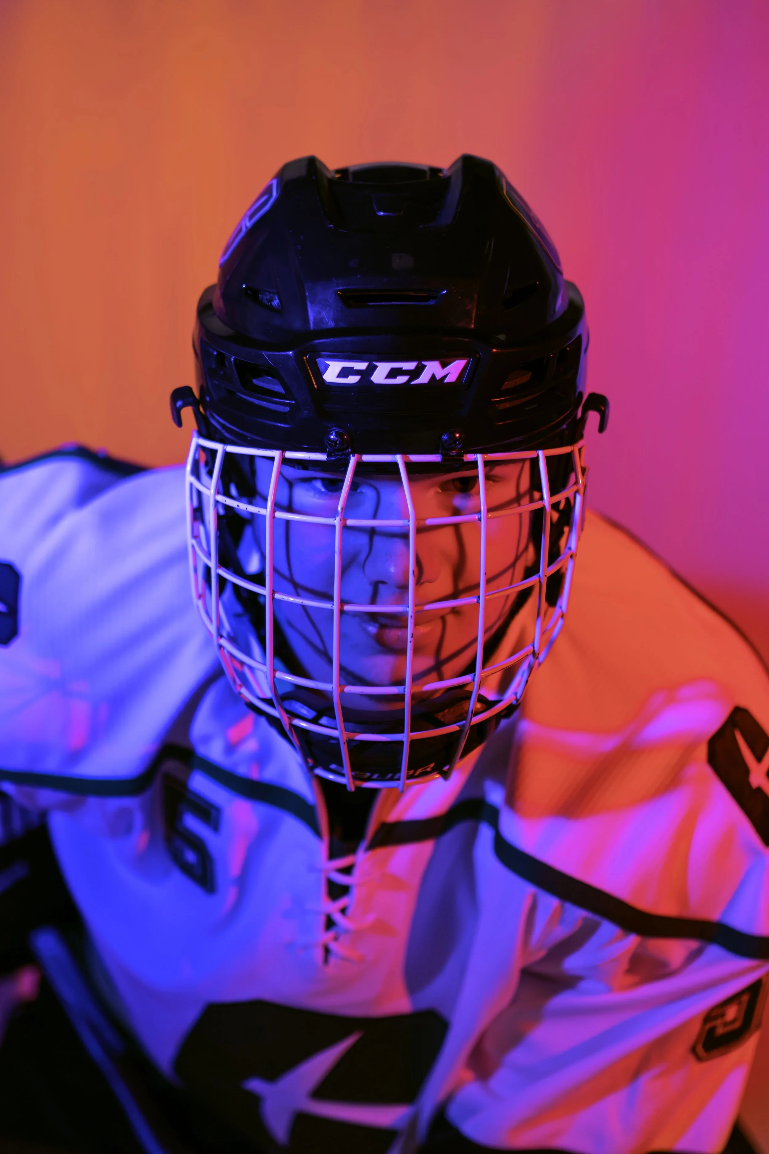 A person in hockey gear wearing a black helmet with a face cage, a white jersey, and looking intensely at the camera, illuminated by vibrant pink and blue lighting.