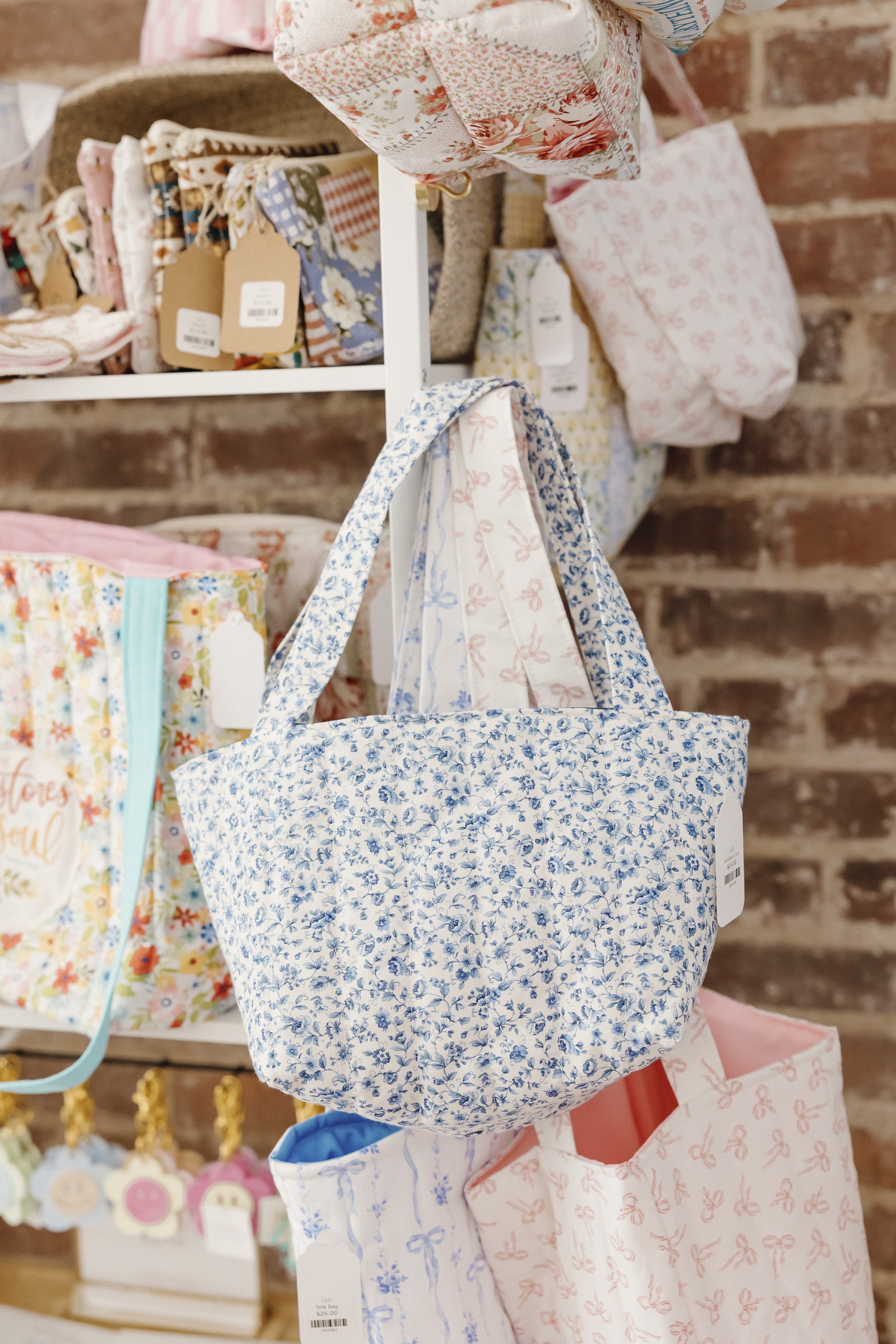 A blue floral fabric tote bag hanging on a display rack in a store, with other fabric gift bags and pouches around it, against a brick wall background.