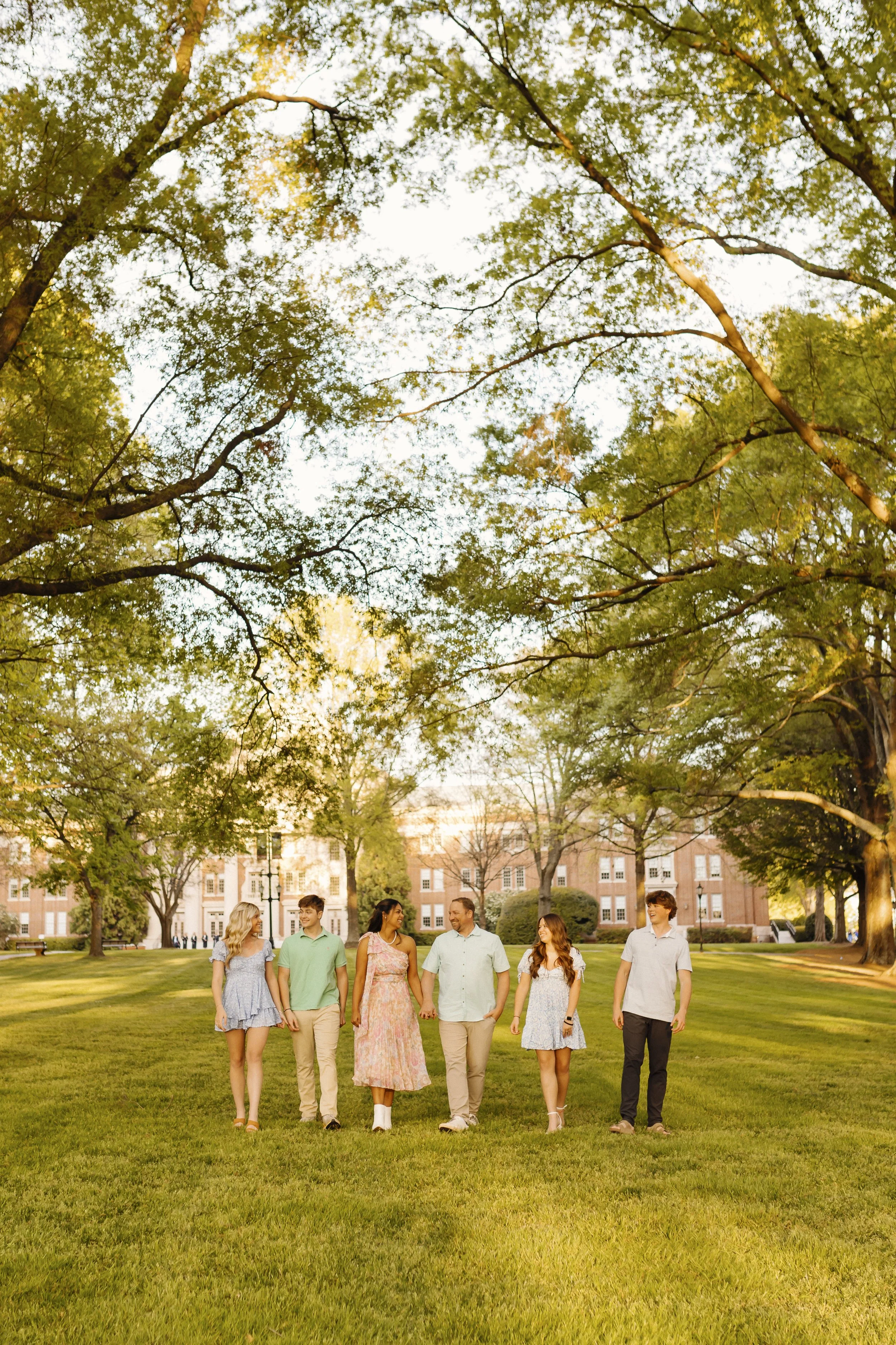 A family walking together in a straight line towards the camera wearing spring pastel colors in a big grass and tree field on Davidson college campus.
