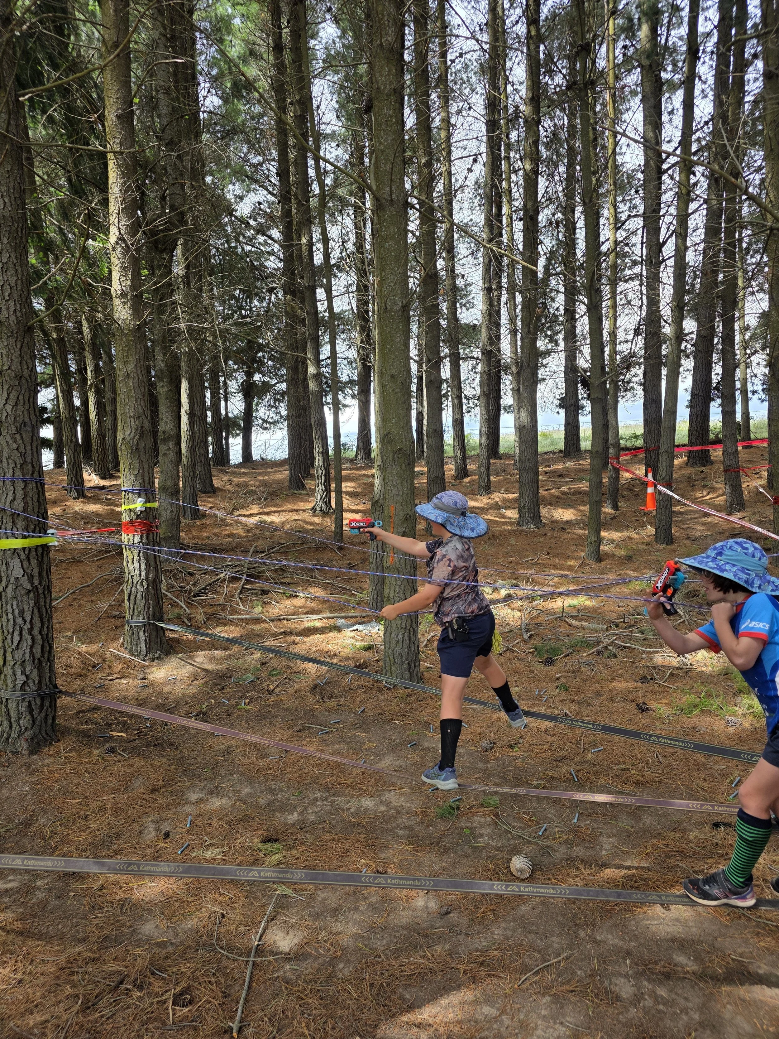 Two children wearing hats and shorts playing or exploring in a wooded forest area with trees and orange and purple tape marking boundaries.