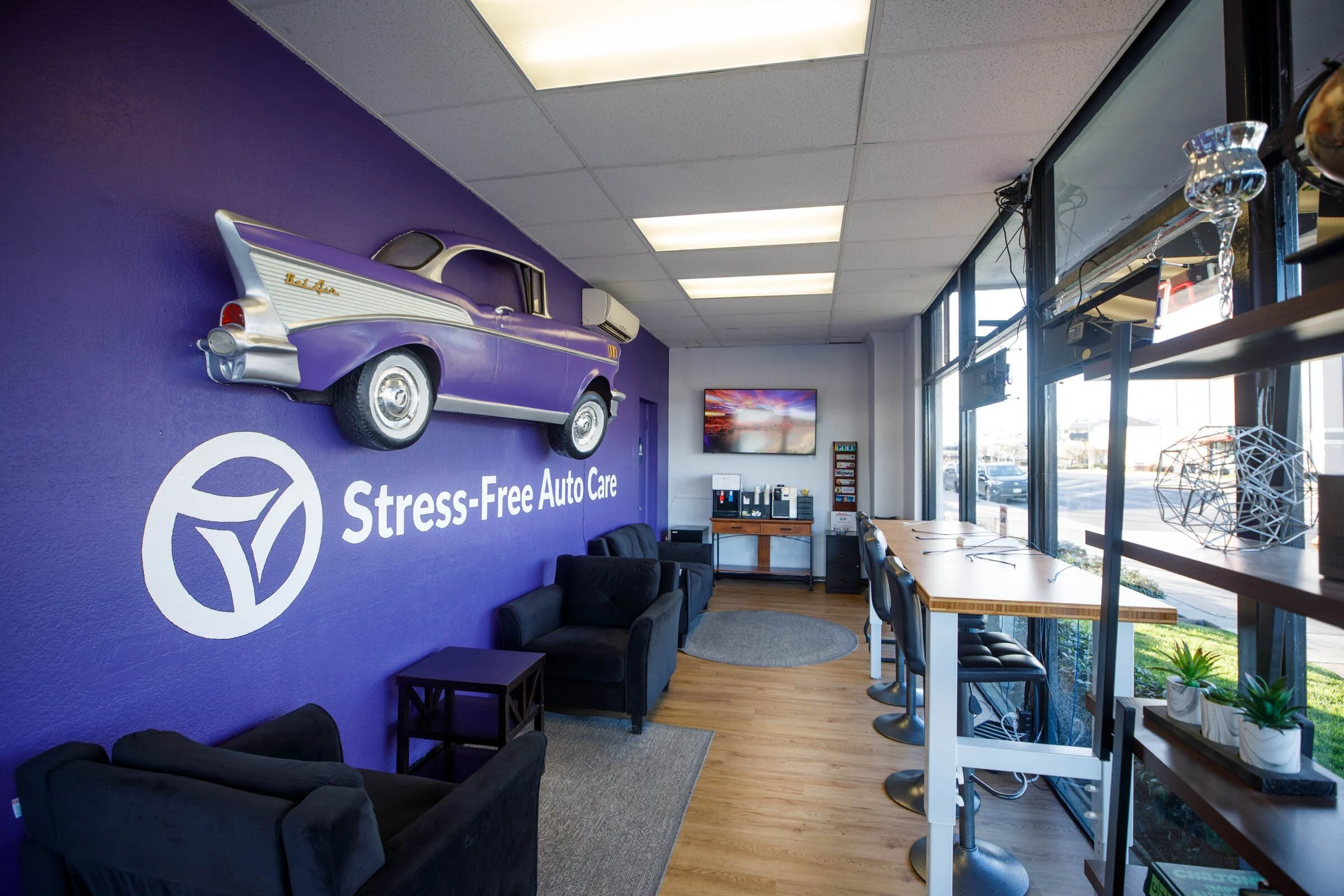 Interior of a stress-free auto care lounge with black sofas, a wooden counter with bar stools, large windows, purple accent wall with a vintage purple car sculpture, and a TV screen.