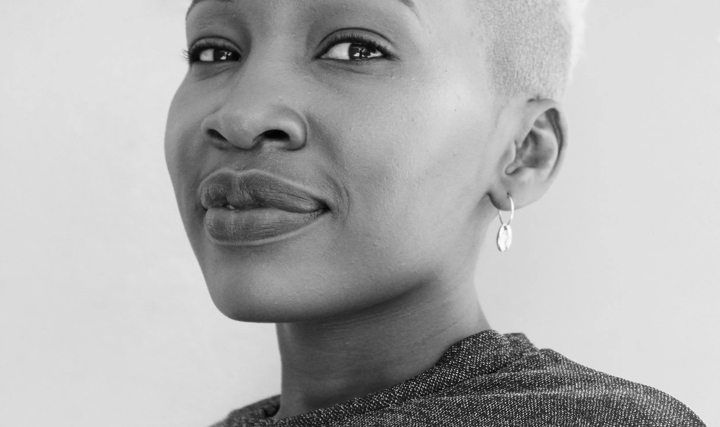Close-up black and white portrait of a woman with short hair, wearing earrings and a textured top, looking at the camera with a subtle smile.