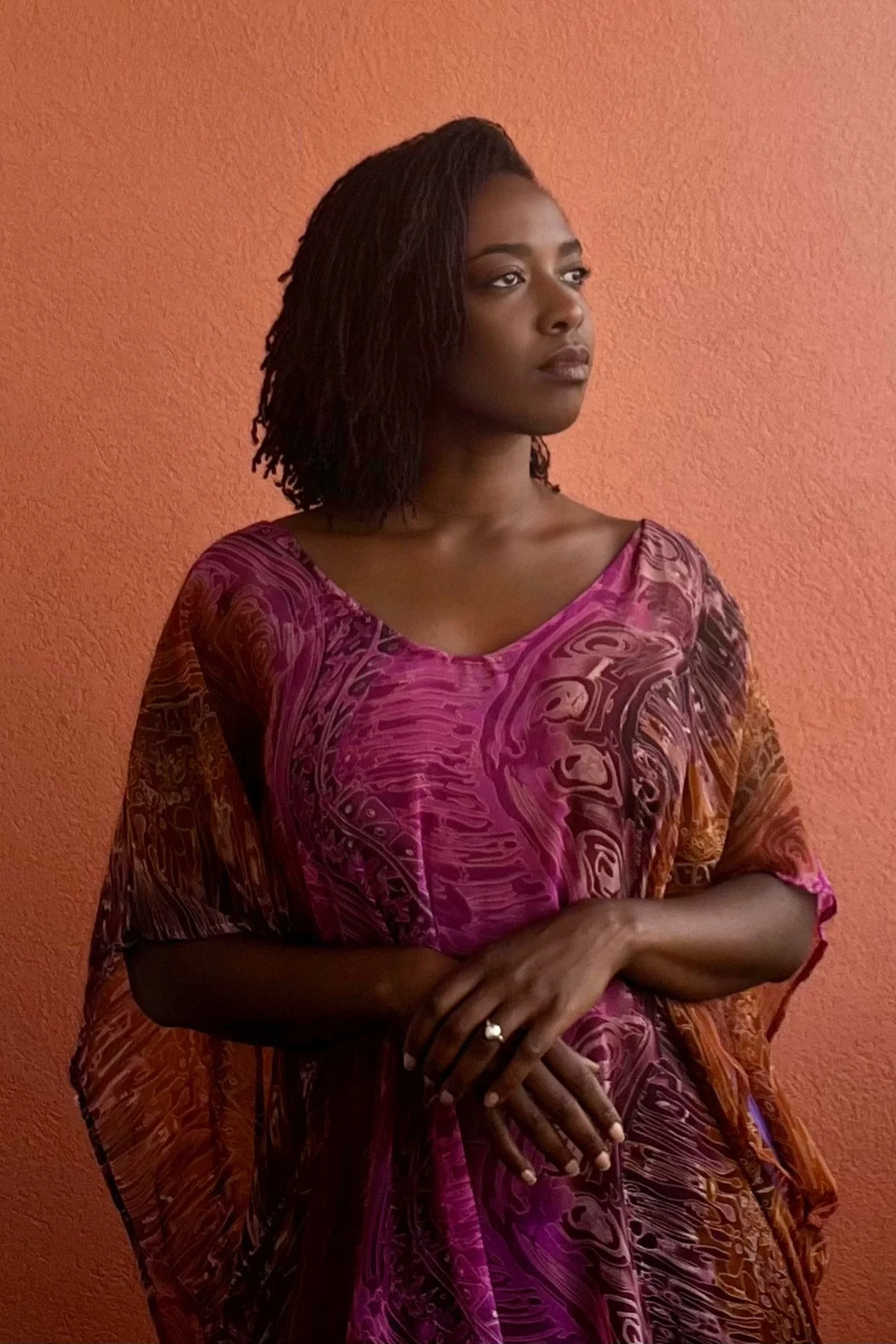 A woman with dark curly hair wearing a colorful, patterned dress standing against a peach-colored wall.