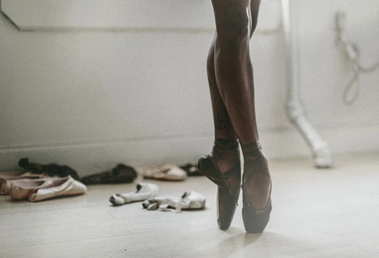 Ballet dancer standing en pointe with shoes on the floor nearby, in a ballet studio.