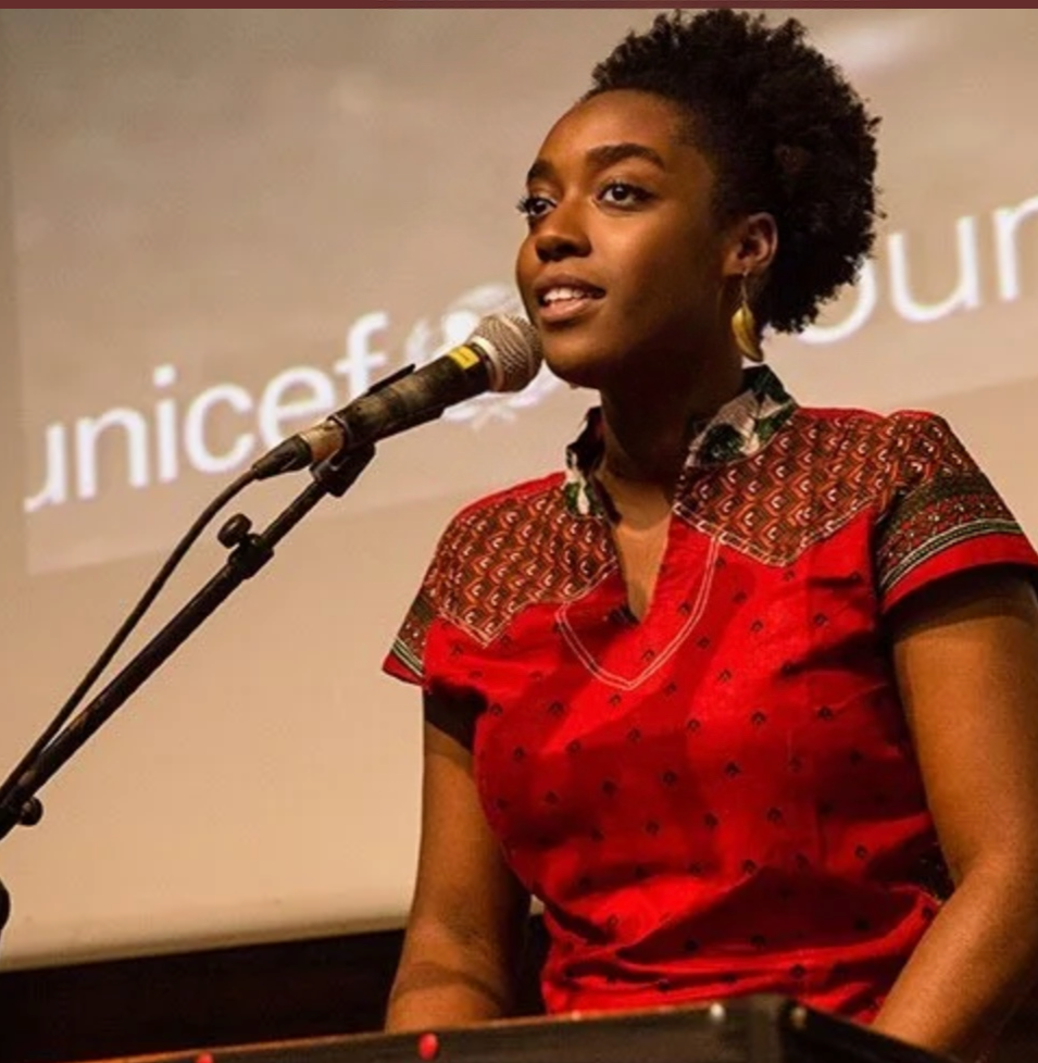 A young woman with short curly hair and brown skin, wearing a red patterned top, is speaking into a microphone on a stand. She appears to be at an event or presentation.