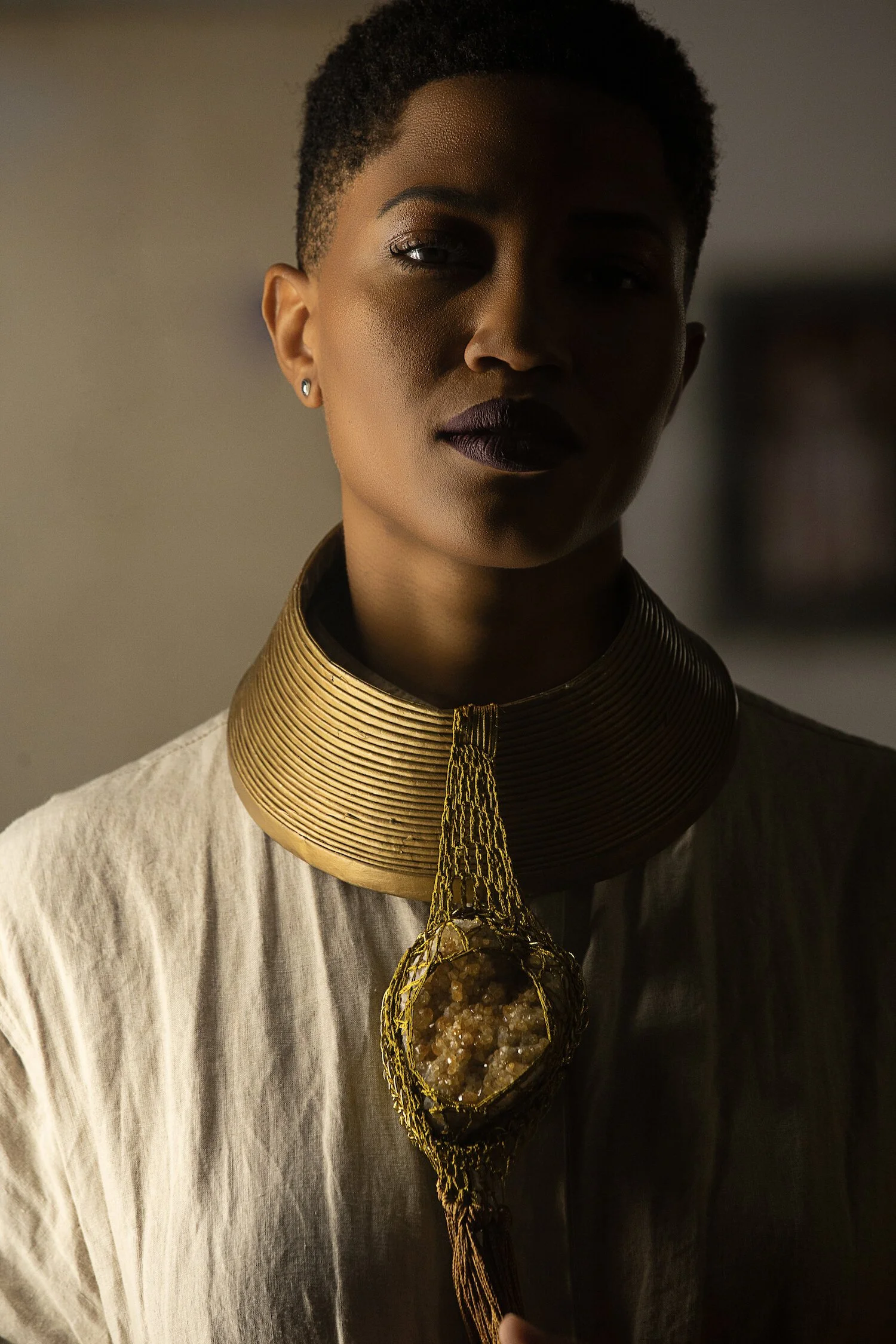 A woman with dark skin and short curly hair wearing dark lipstick, with a large, gold, circular necklace featuring a crystal pendant around her neck, dressed in a light-colored, loose-fitting top.