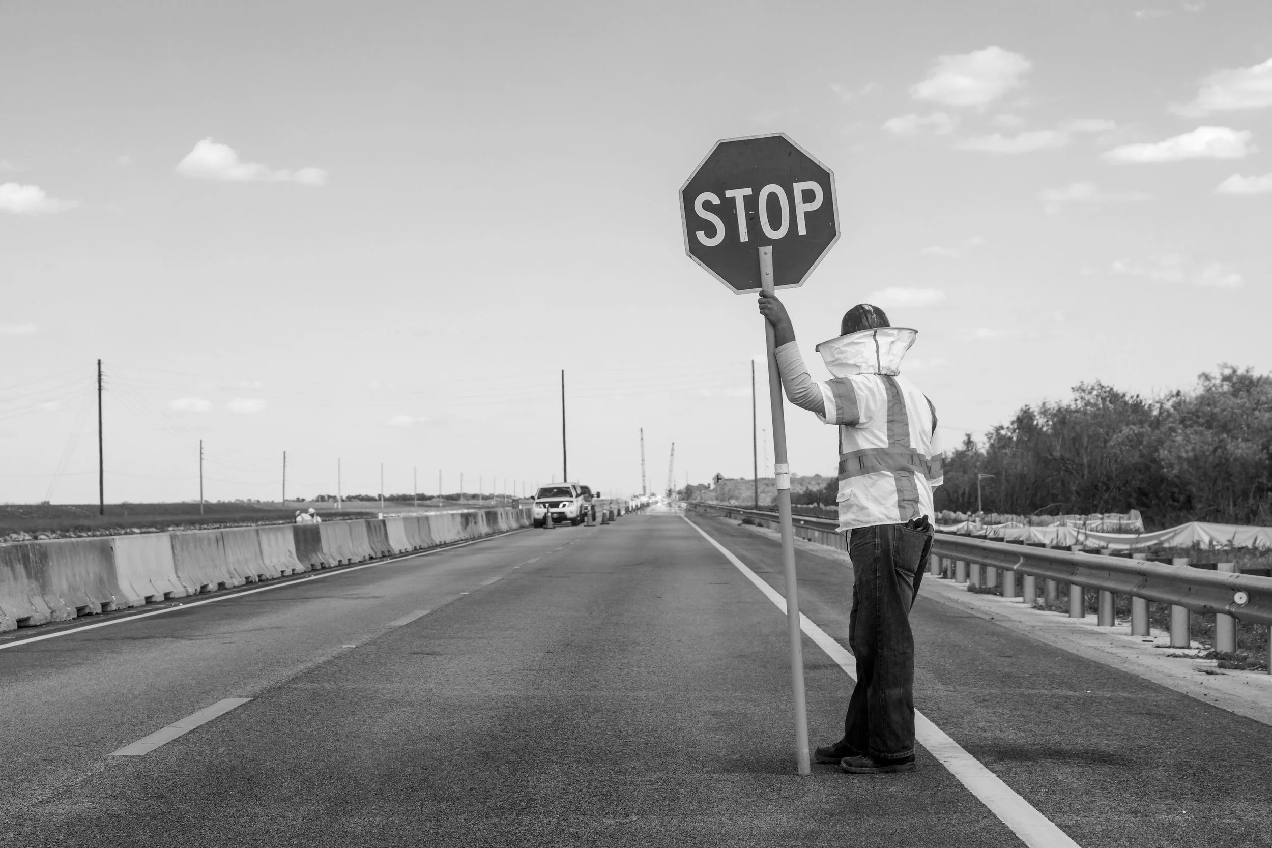 A person wearing a reflective safety vest and dark jeans holding a stop sign on an empty road with traffic cones and a few cars in the distance under a partly cloudy sky.