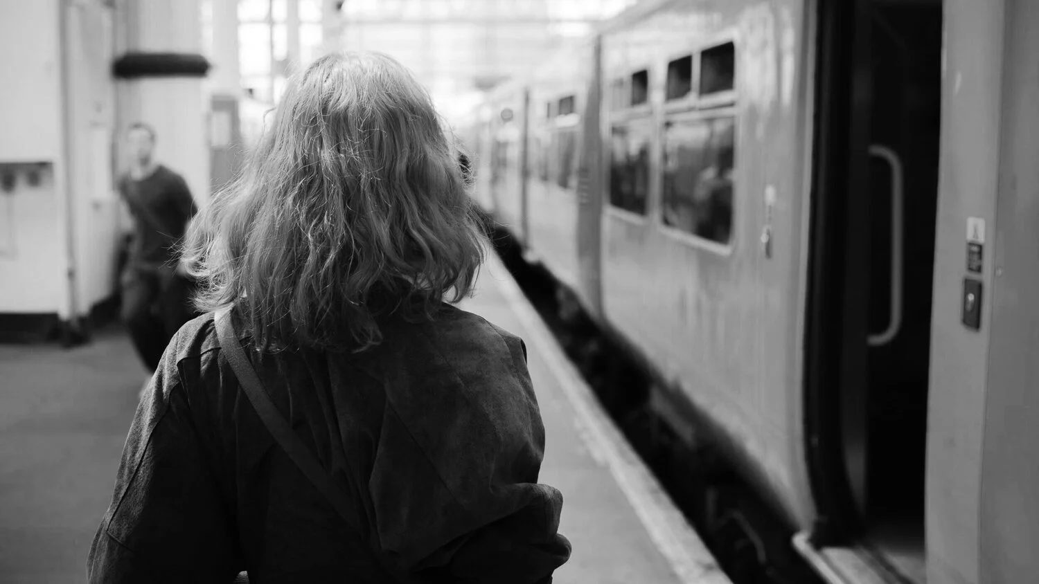 A woman with shoulder-length hair is standing on a subway platform, looking at a train. The image is in black and white.
