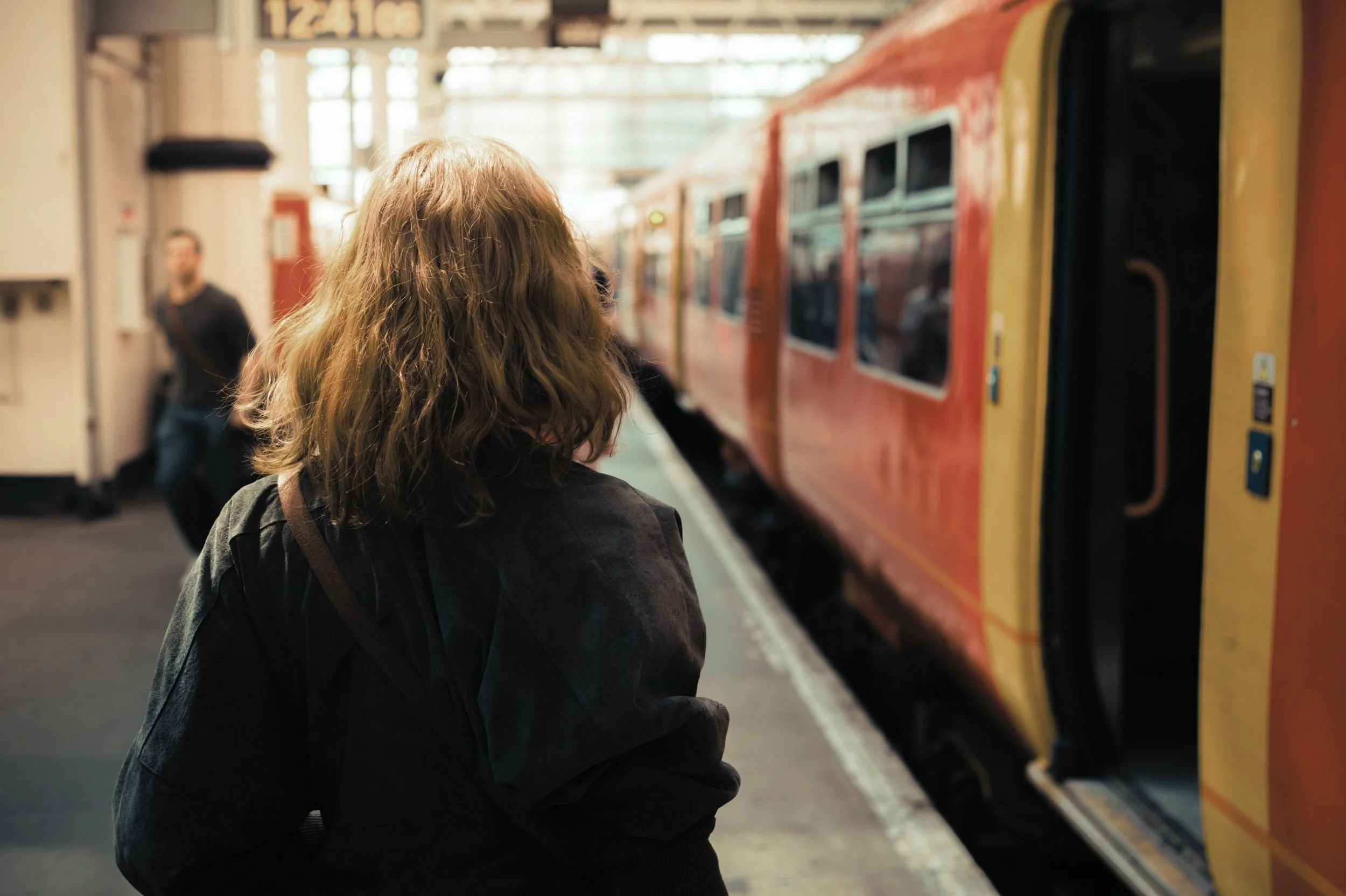 A woman with shoulder-length brown hair, dressed in black, stands on a train platform waiting for a red and yellow train to arrive, with a man walking in the background.