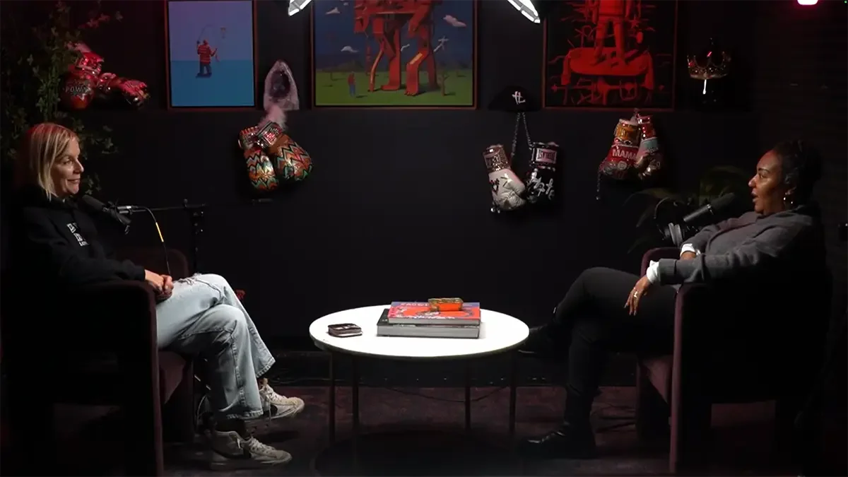 Keri and Achea are seated in armchairs facing each other in a podcast studio. They are wearing headphones and speaking into microphones. The background is decorated with holiday stockings, framed cartoon artwork, and festive decorations.