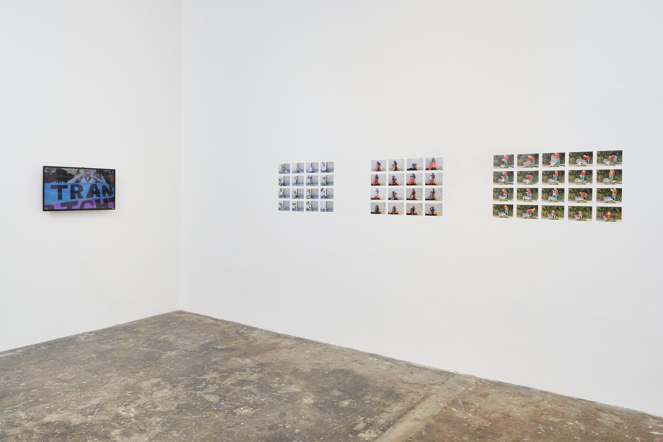 Photo of an art exhibition featuring photographs of a person in various poses and outfits, displayed on a gallery white wall, with a small television screen on the left showing text.