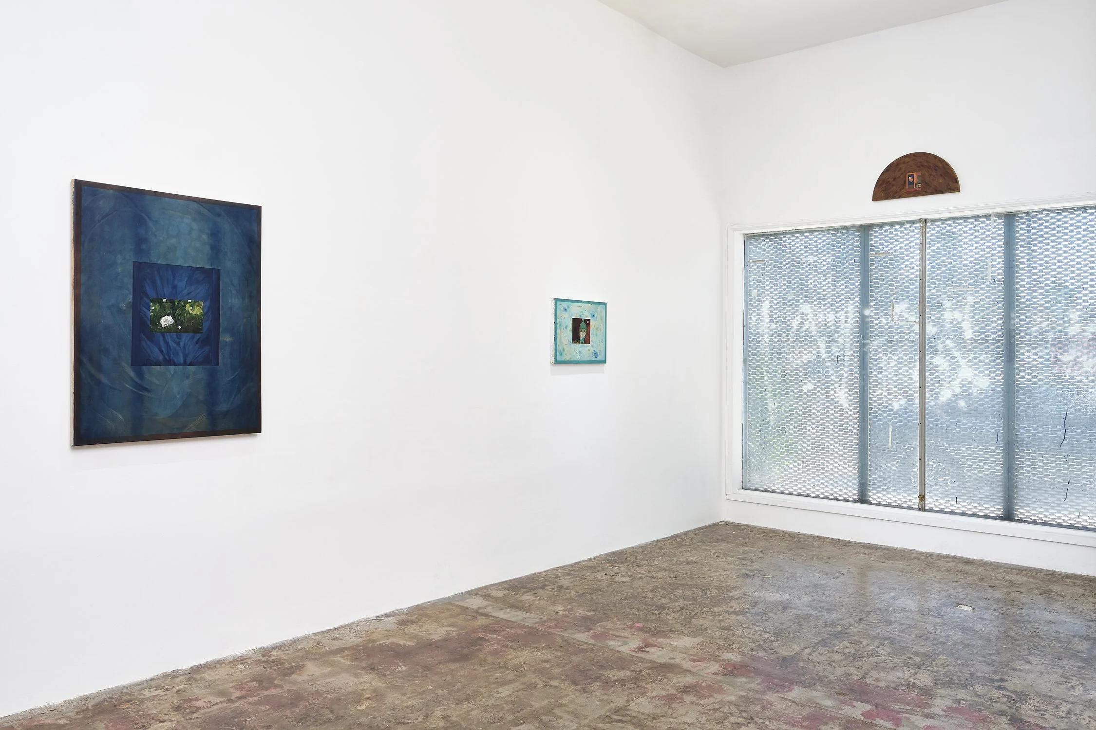 Interior of an art gallery with three paintings on white walls and a large window with metal security screen.