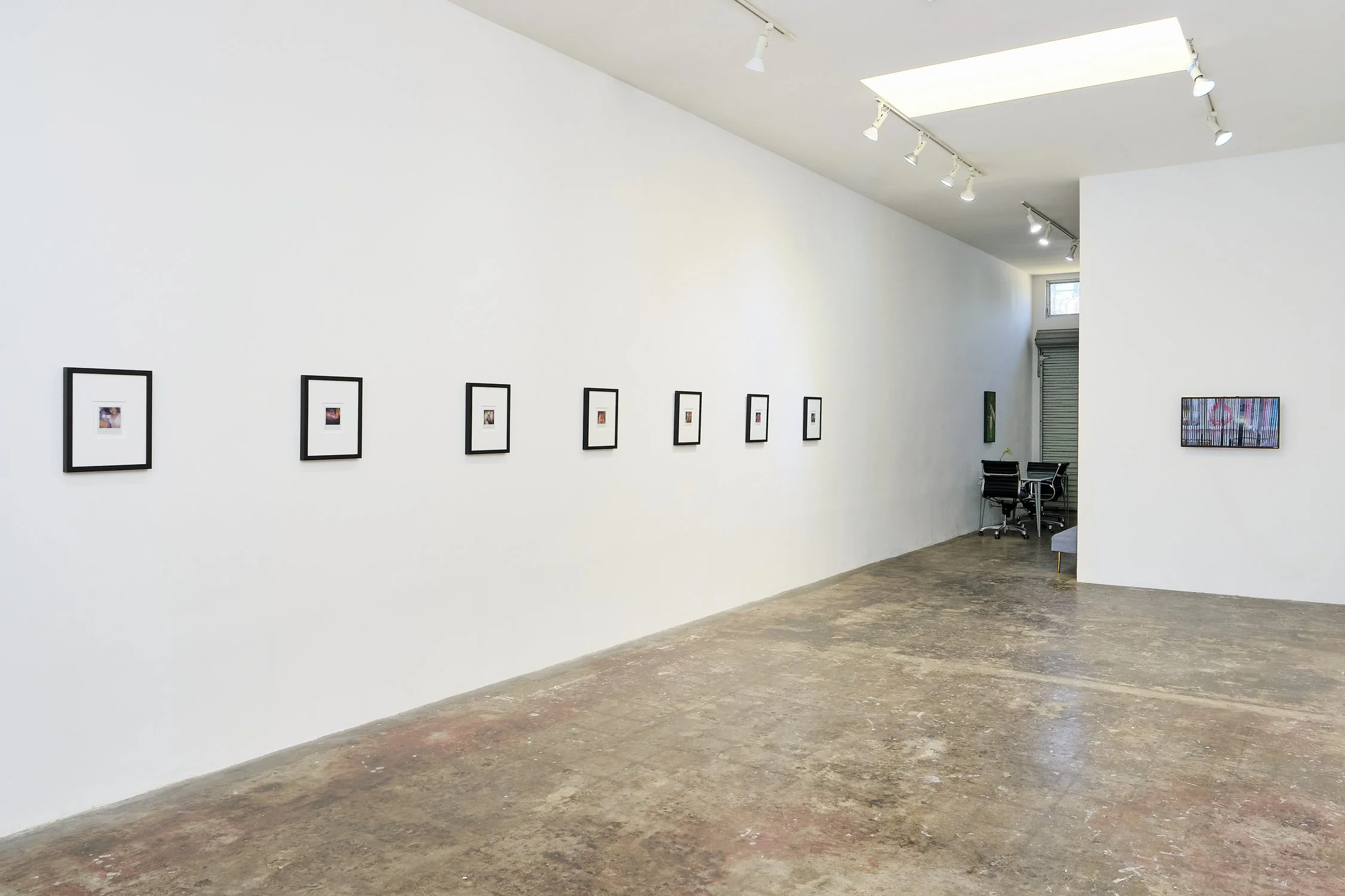 An art gallery with a white wall displaying framed photographs, a corner with a table and chairs, and a small window near the ceiling letting in natural light.