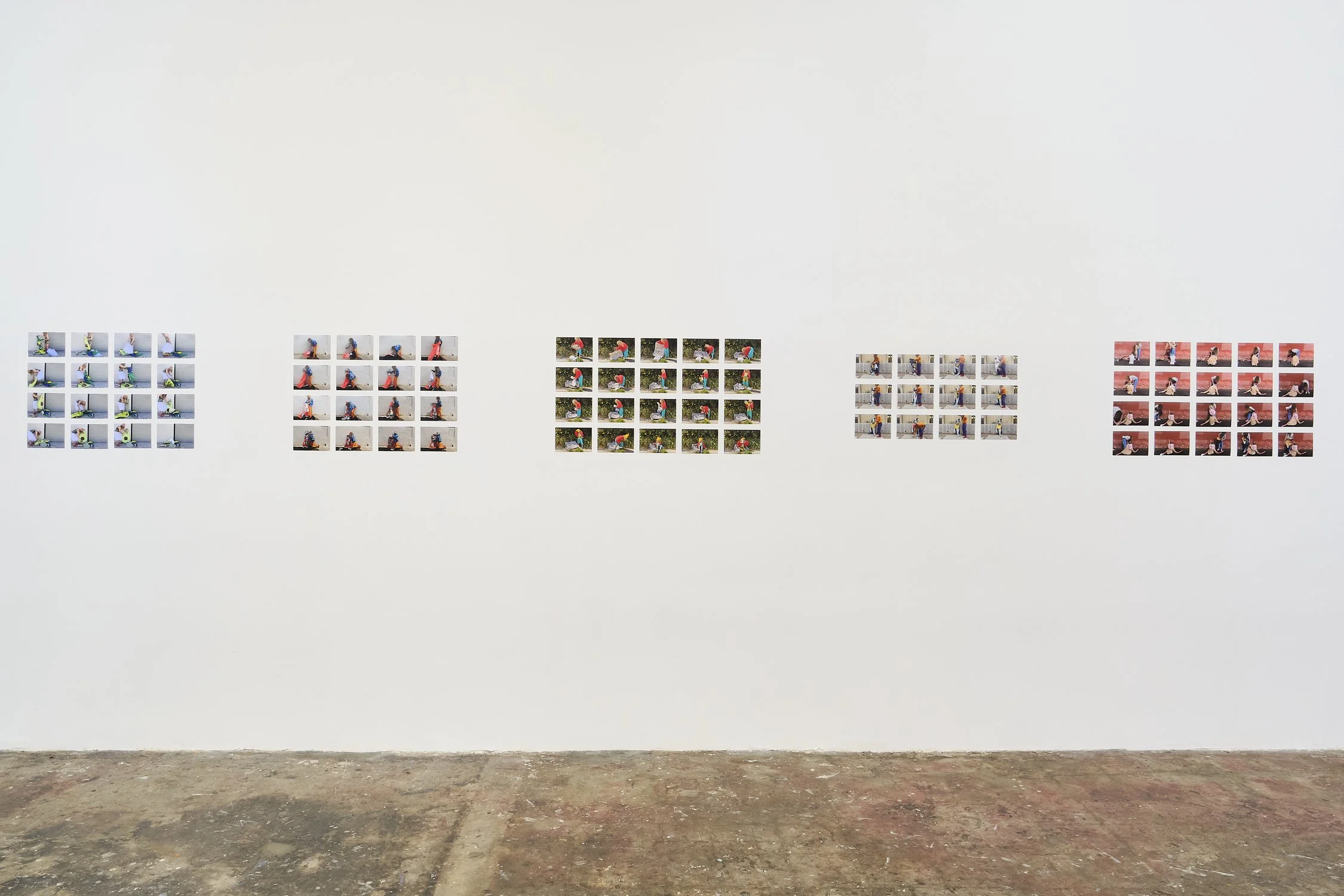 A gallery of small photographs arranged on a white wall with a concrete floor beneath.