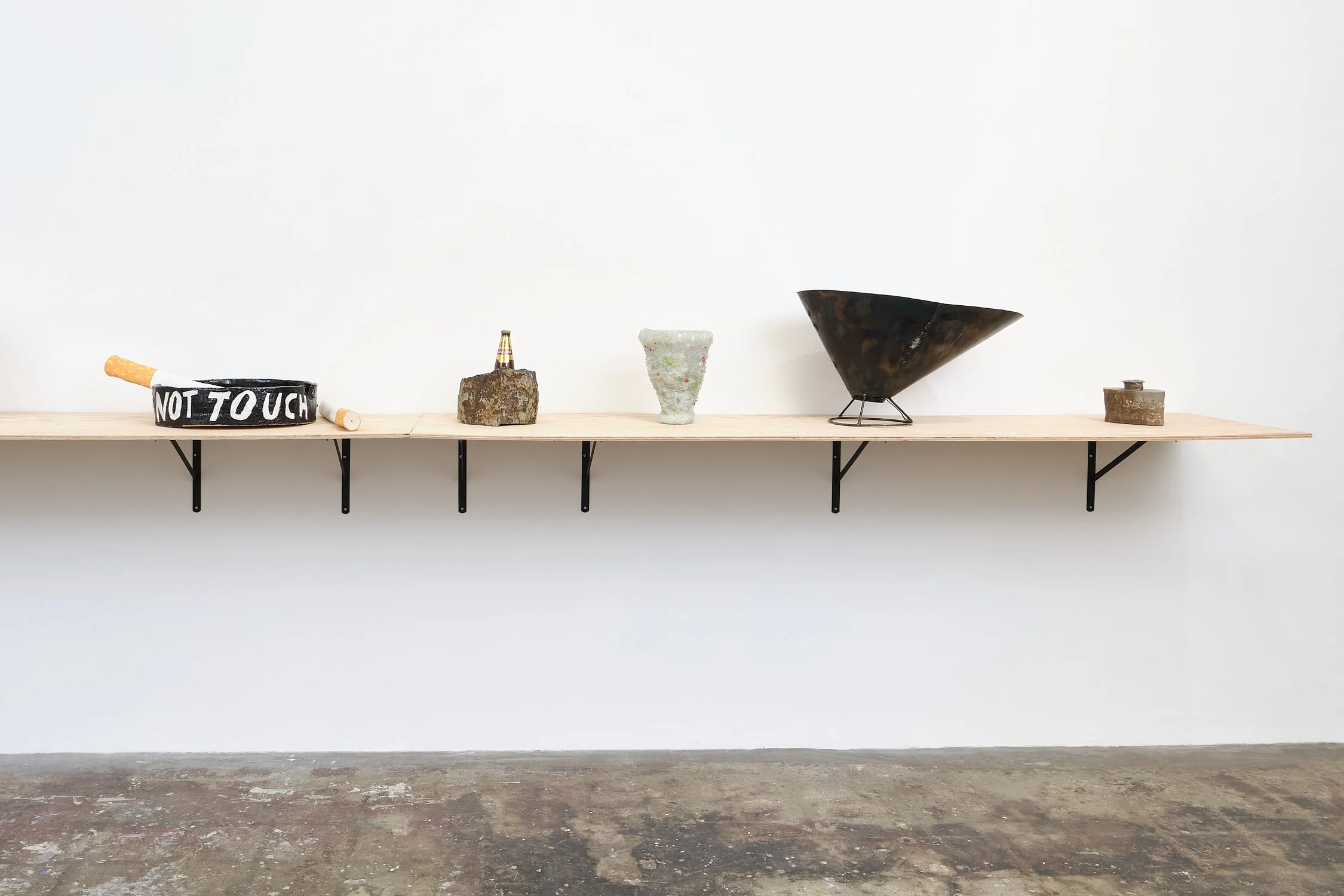 A minimalist wooden shelf mounted on a wall with various objects displayed, including a roll of tape with 'NOT TOUCH' written on it, a small bottle on a rock, a ceramic vase, a metallic cone-shaped sculpture, and a small wooden box.