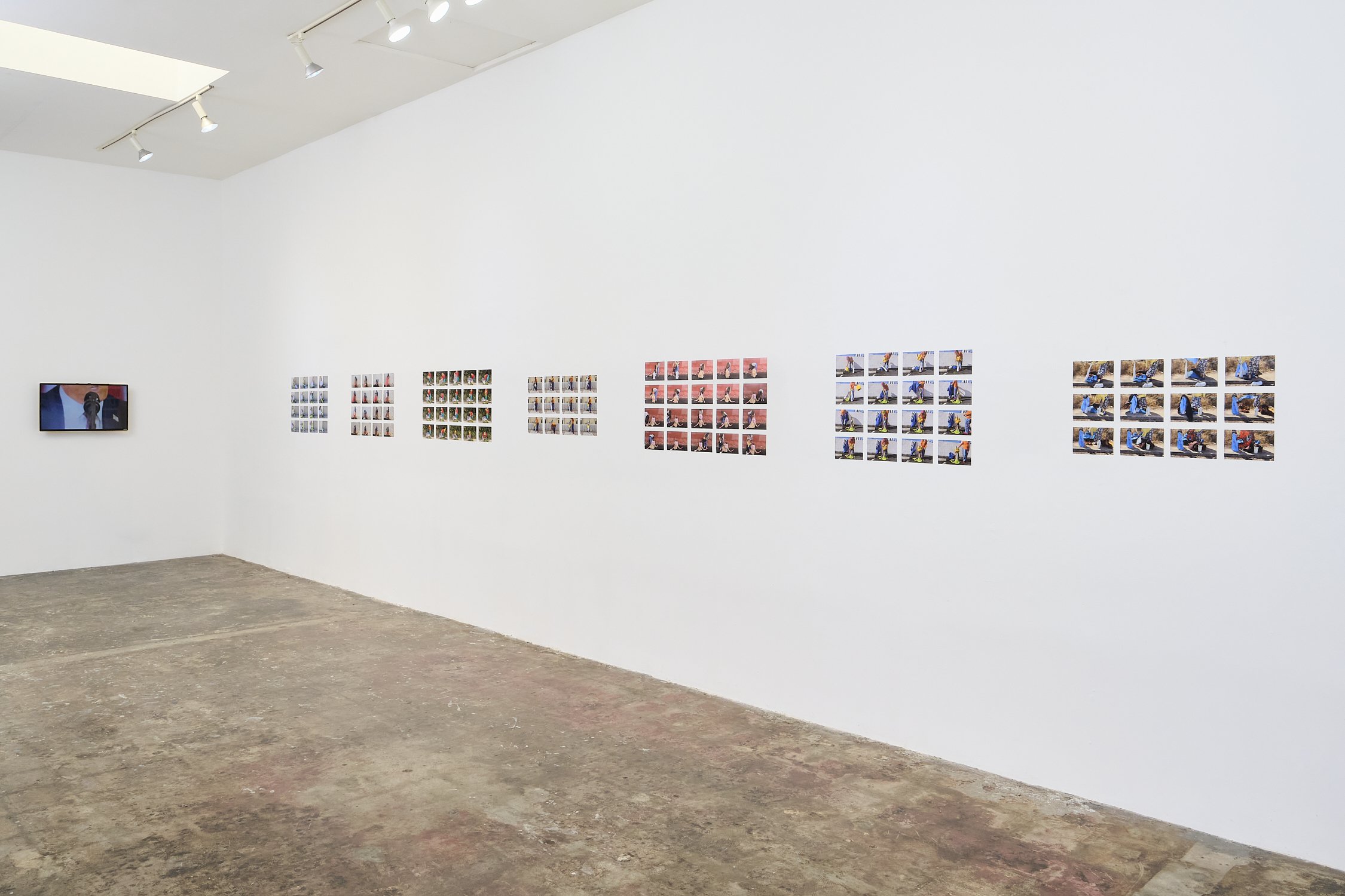 Gallery wall with multiple small photographs arranged in rows, alongside a video monitor displaying a person speaking, in an art gallery with white walls and a concrete floor.