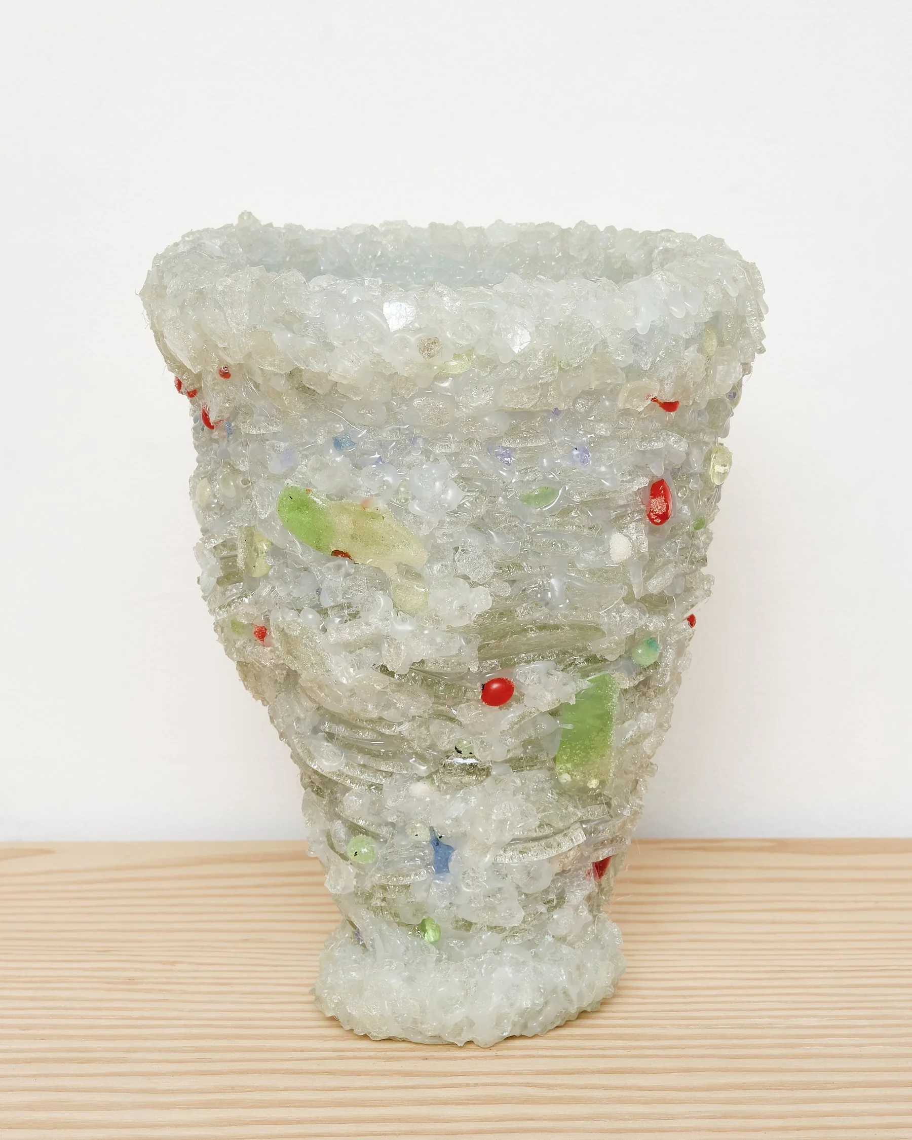 A sculpture resembling a tall, tapered vase made of translucent, textured material with colorful embedded fragments, placed on a wooden surface against a white background.