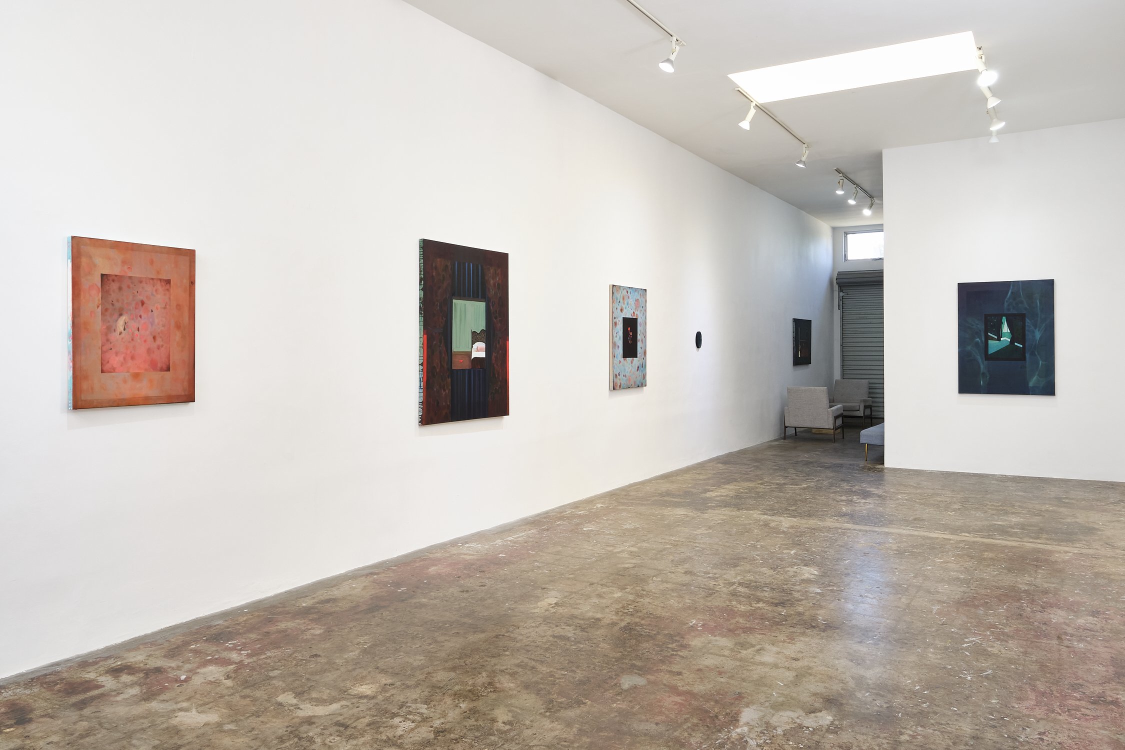An art gallery with a white wall displaying abstract paintings, a concrete floor, and seating area with chairs near the back.