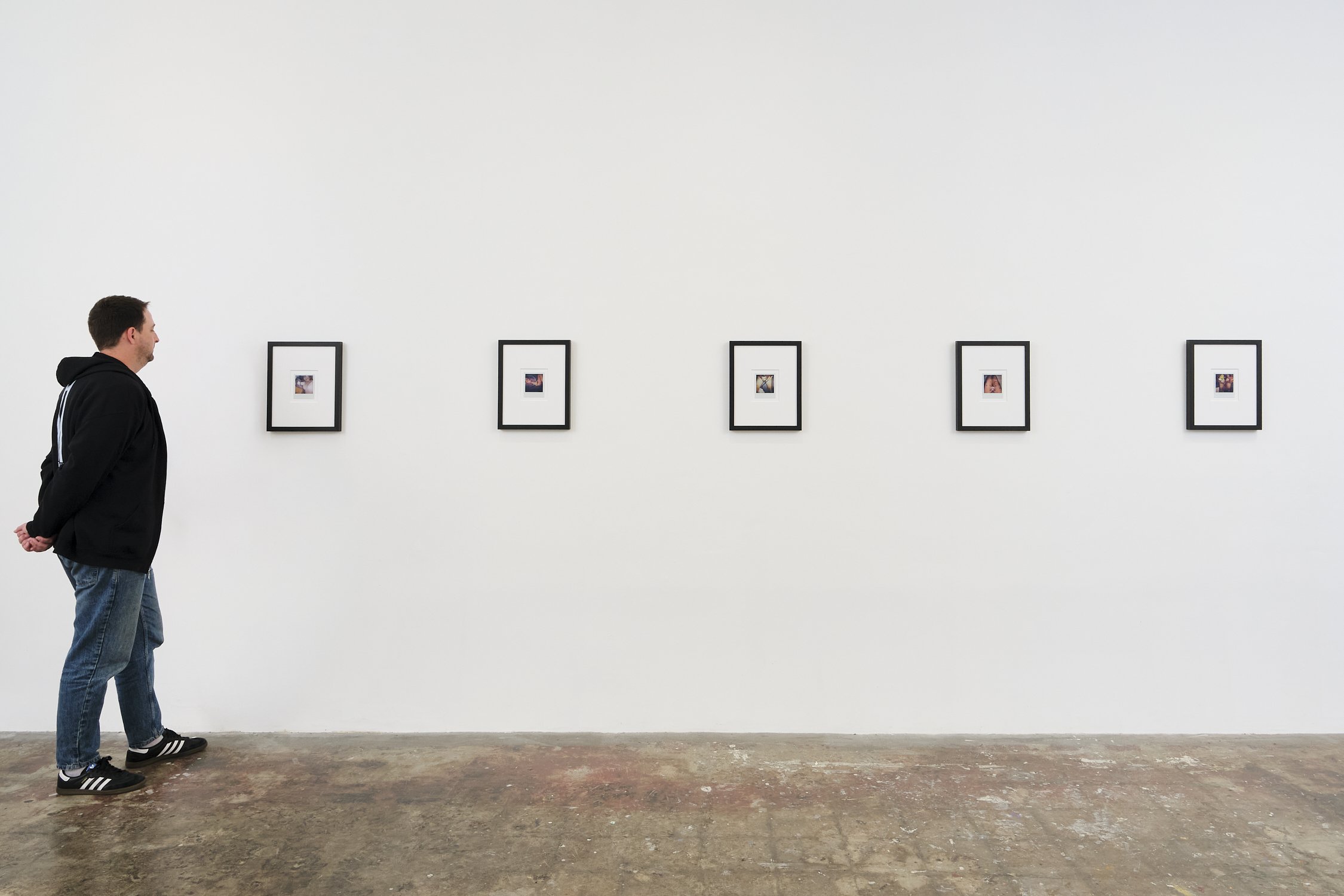A man with short dark hair, wearing a black hoodie, jeans, and sneakers, stands facing a white gallery wall with five framed photographs. The gallery wall is plain and minimalist, and the man appears to be viewing the artwork.