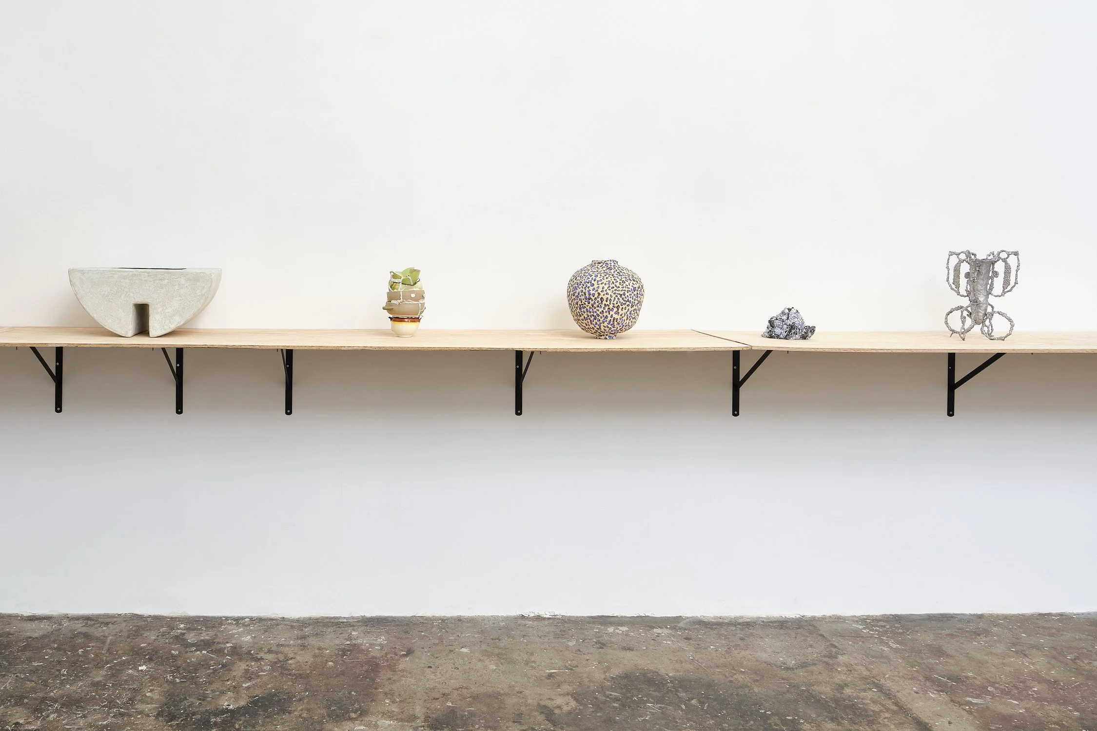A minimalist display shelf with five decorative items: a half-circle stone sculpture, a small stack of colorful ceramic bowls, a round multicolored mosaic vase, a small gray rock, and a target-shaped metal sculpture. The shelf is mounted on a white w