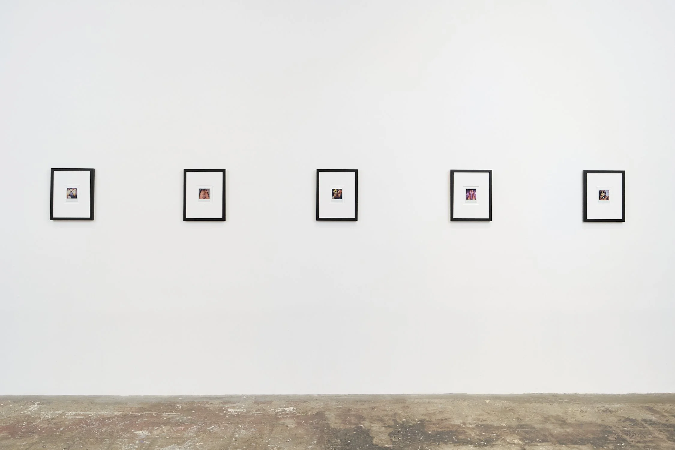 An art gallery with five framed photographs hanging on a white wall. The gallery has a concrete floor and no visible visitors.