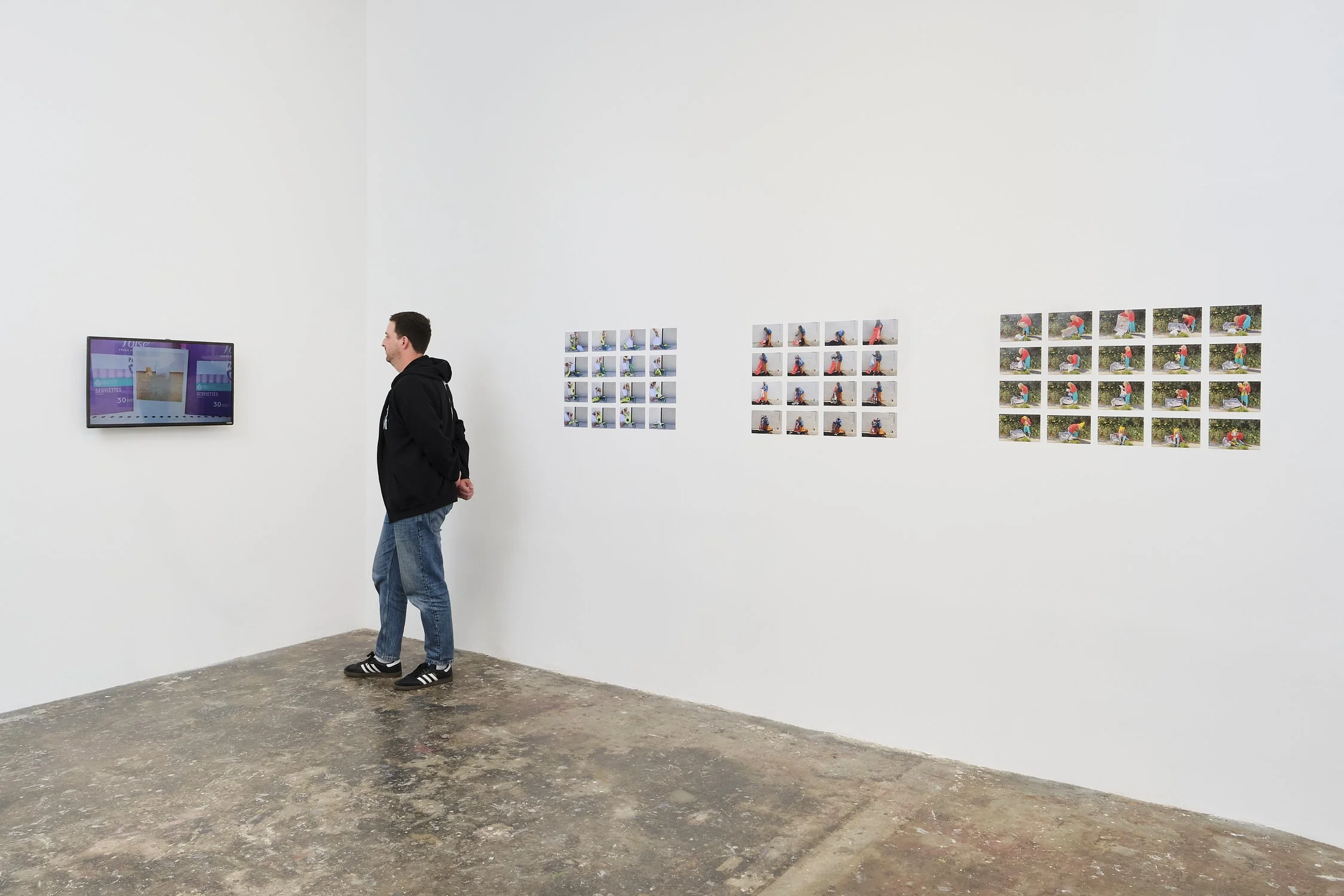 A man in a black hoodie and jeans stands in an art gallery, looking at a video display on the white wall. To his right, there are three sets of photographs arranged in rows, depicting a person in colorful clothing performing outdoor activities.