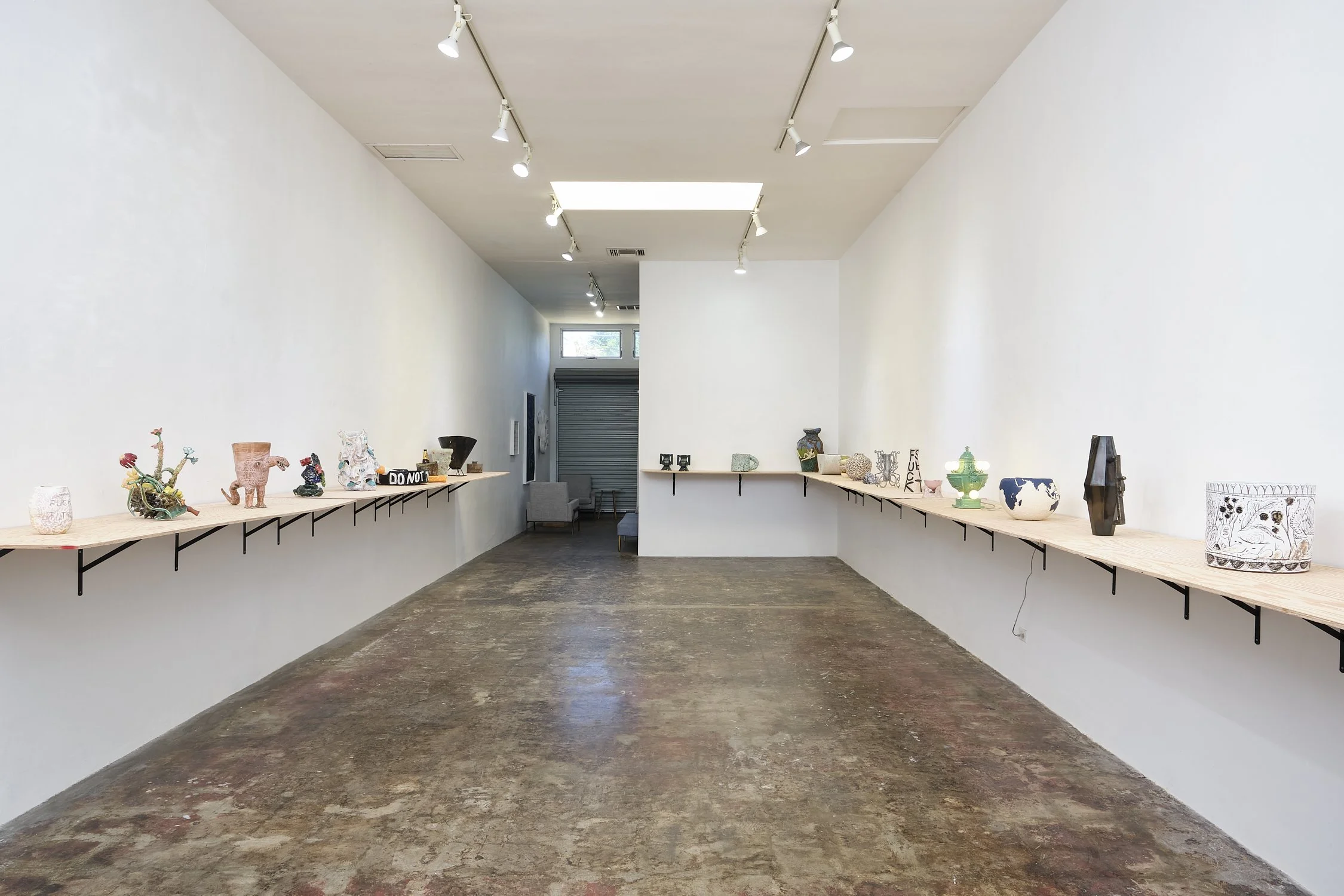 Art gallery with ceramic and pottery sculptures displayed on long wooden shelves along white walls, with track lighting overhead.