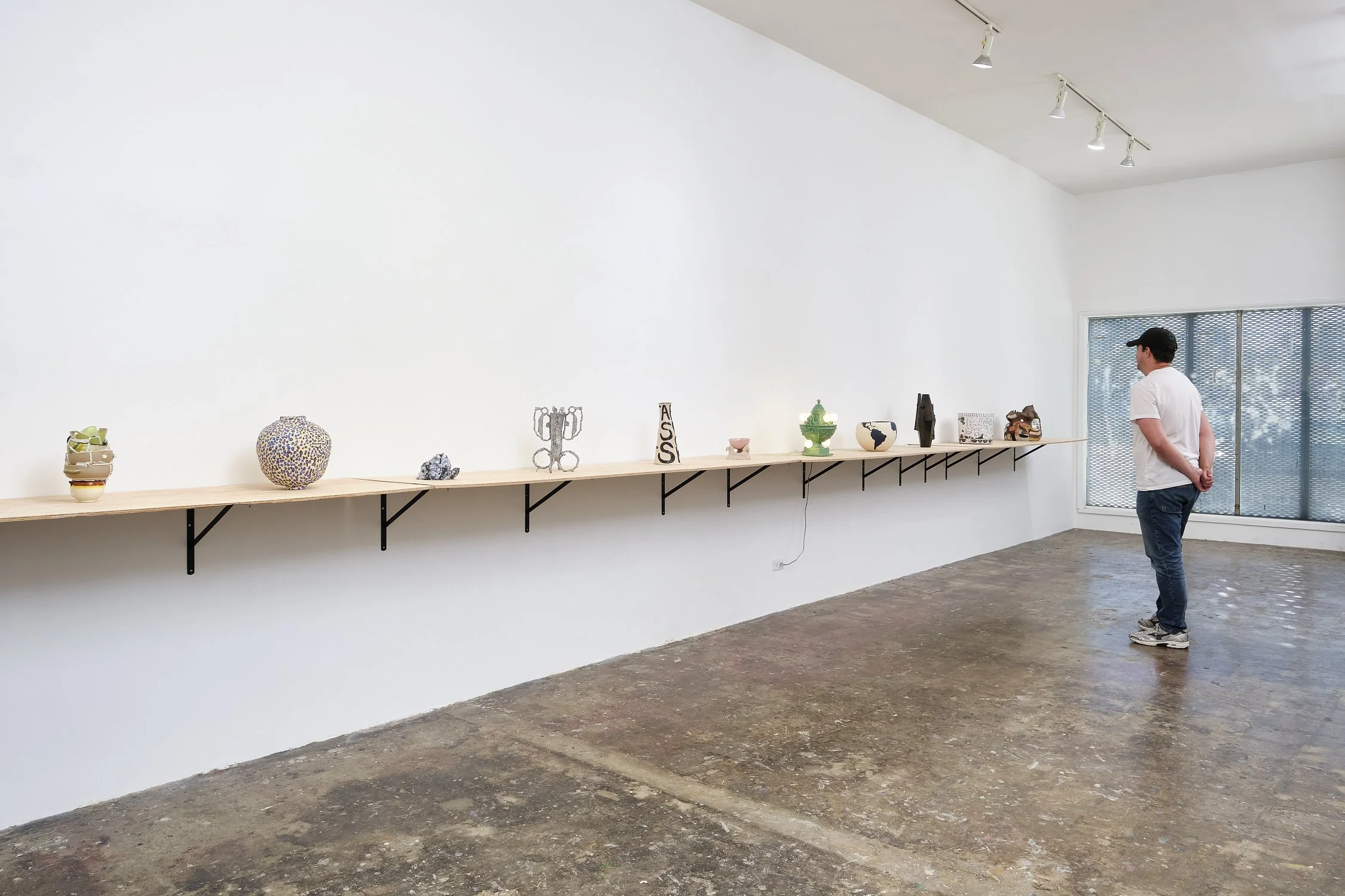 A man viewing a collection of ceramic art pieces displayed on a long wooden shelf in an art gallery with white walls and a concrete floor.