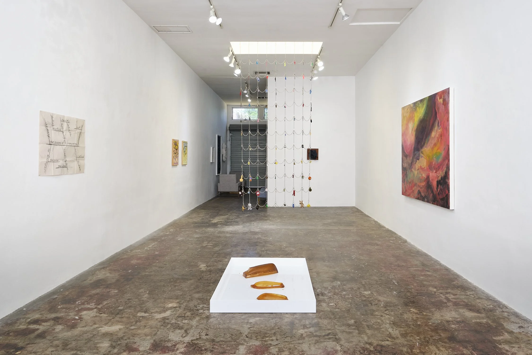 An art gallery with white walls and concrete floors displaying various paintings and sculptures, including a suspended chain installation at the center and three organisms on a white pedestal in the foreground.