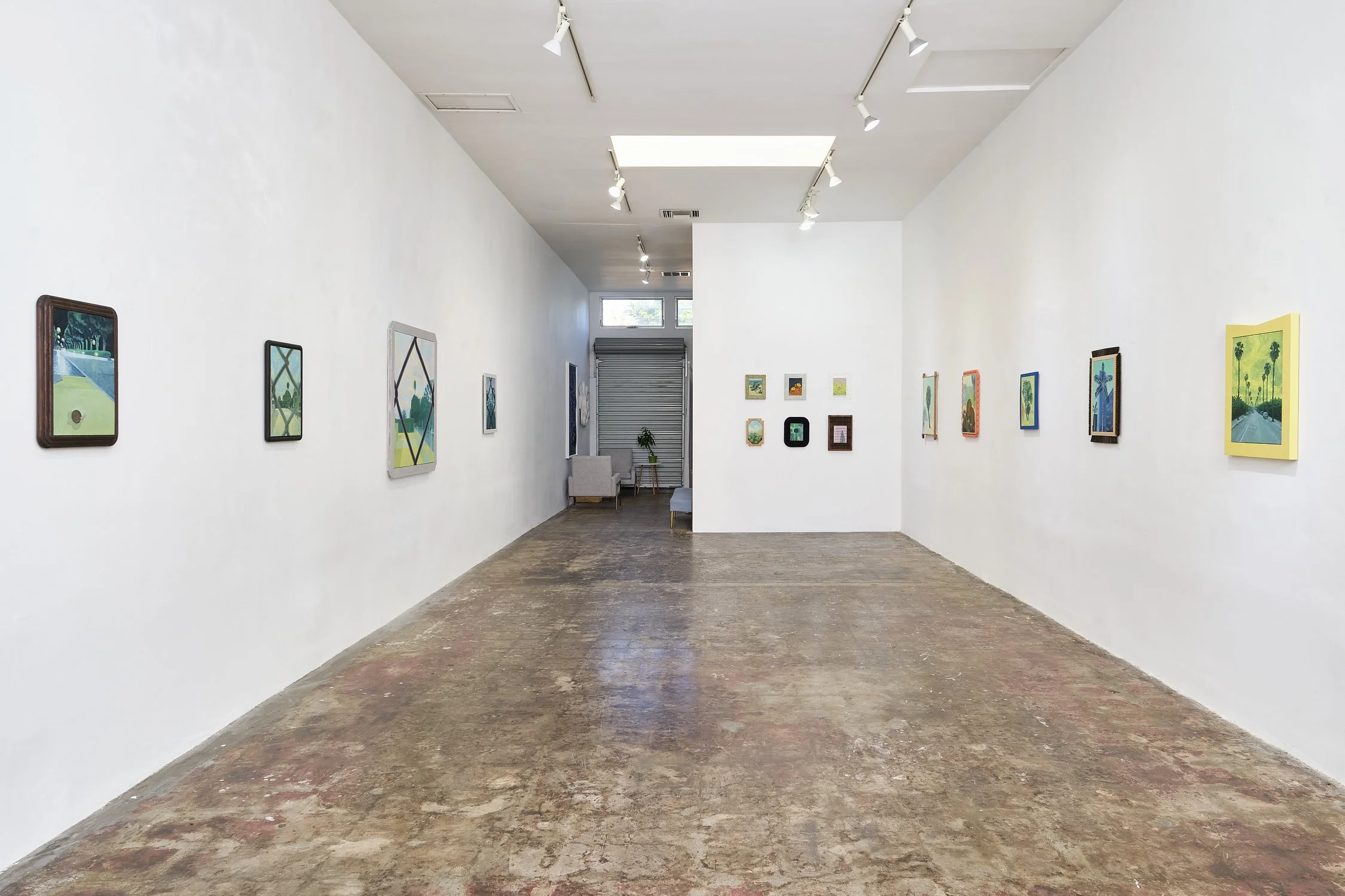 An art gallery with white walls displaying various colorful framed artworks, a polished concrete floor, track lighting on the ceiling, and a seating area with a gray couch and chairs near the back.