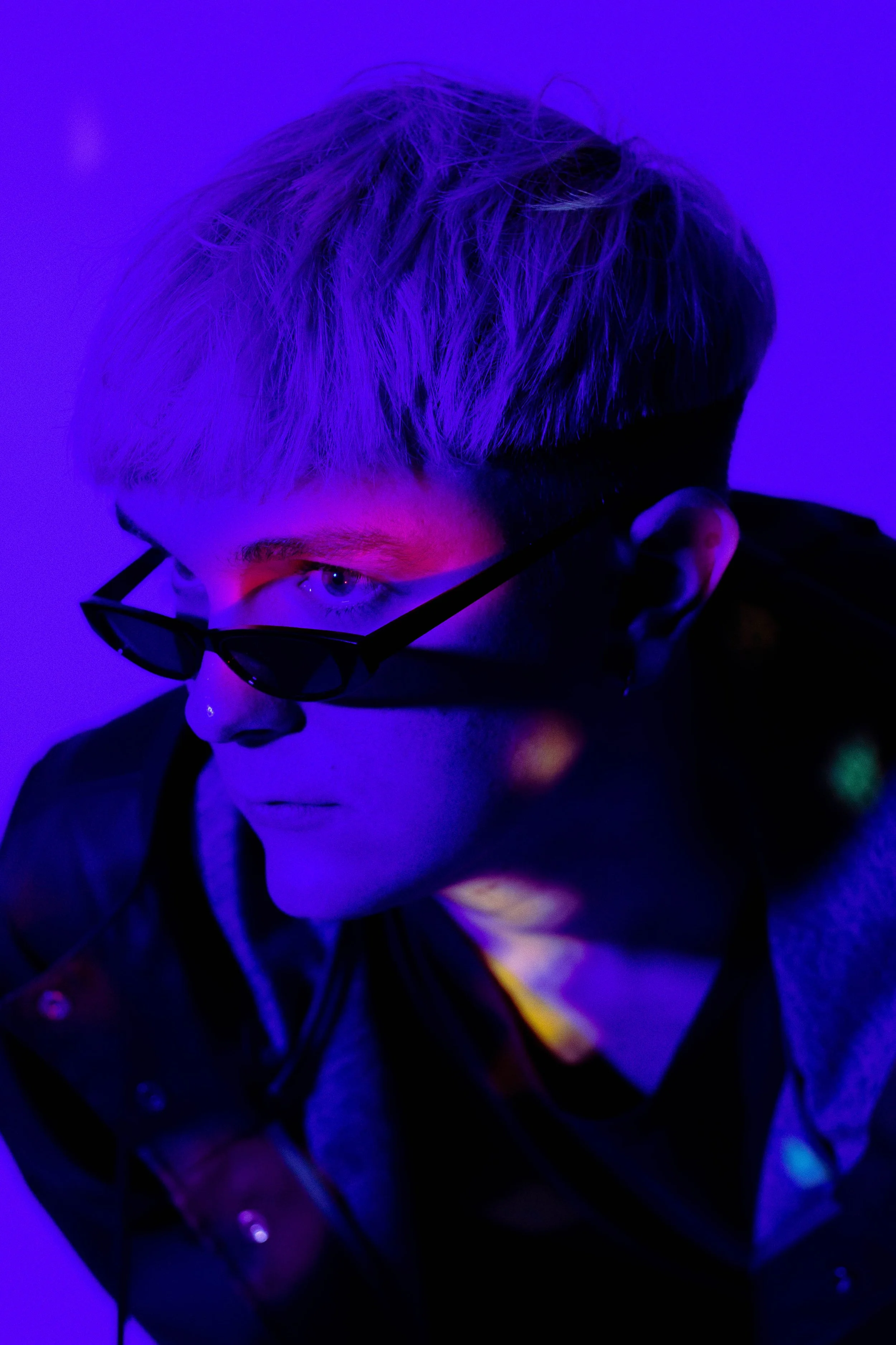 A person with blond hair wearing black-rimmed glasses, illuminated by vibrant purple and blue lighting, looking downward with a serious expression.