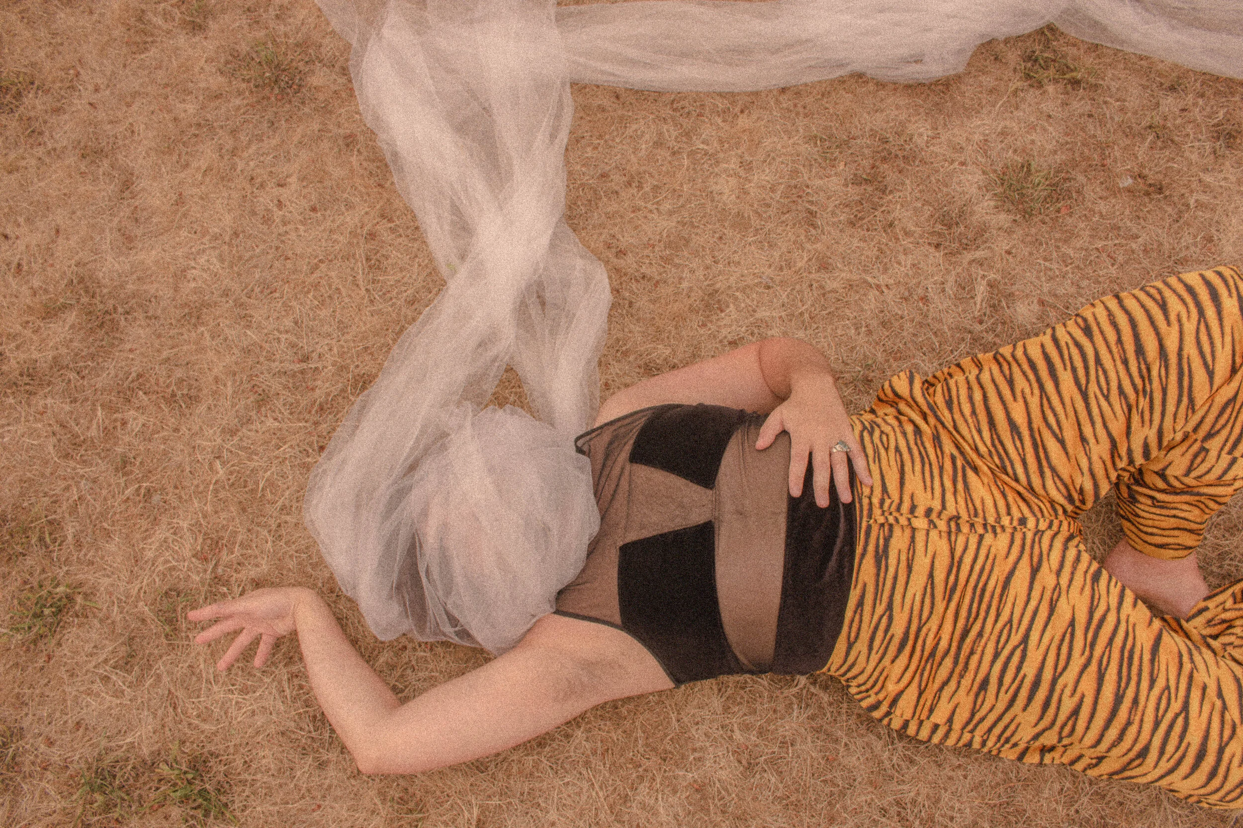 A person lying on dry grass, wearing a black top and beige pants with a tiger-print shirt. The person's face is not visible, and they are posed with one hand resting on their stomach and the other arm extended on the ground.