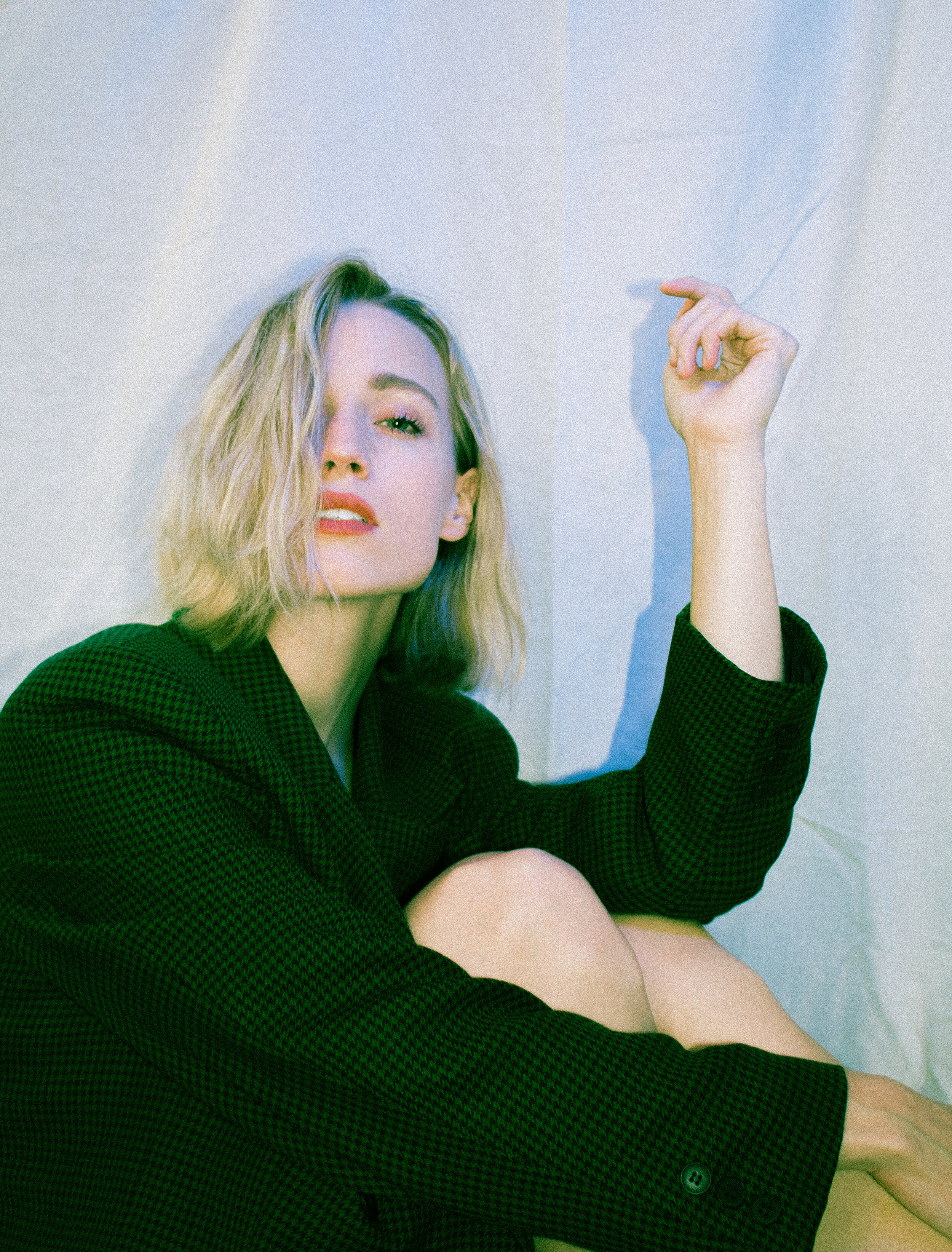 A woman with blonde hair is sitting against a white wall, wearing a dark textured blazer, partially covering her face with her hair, and resting her arm on her knee while holding a cigarette.