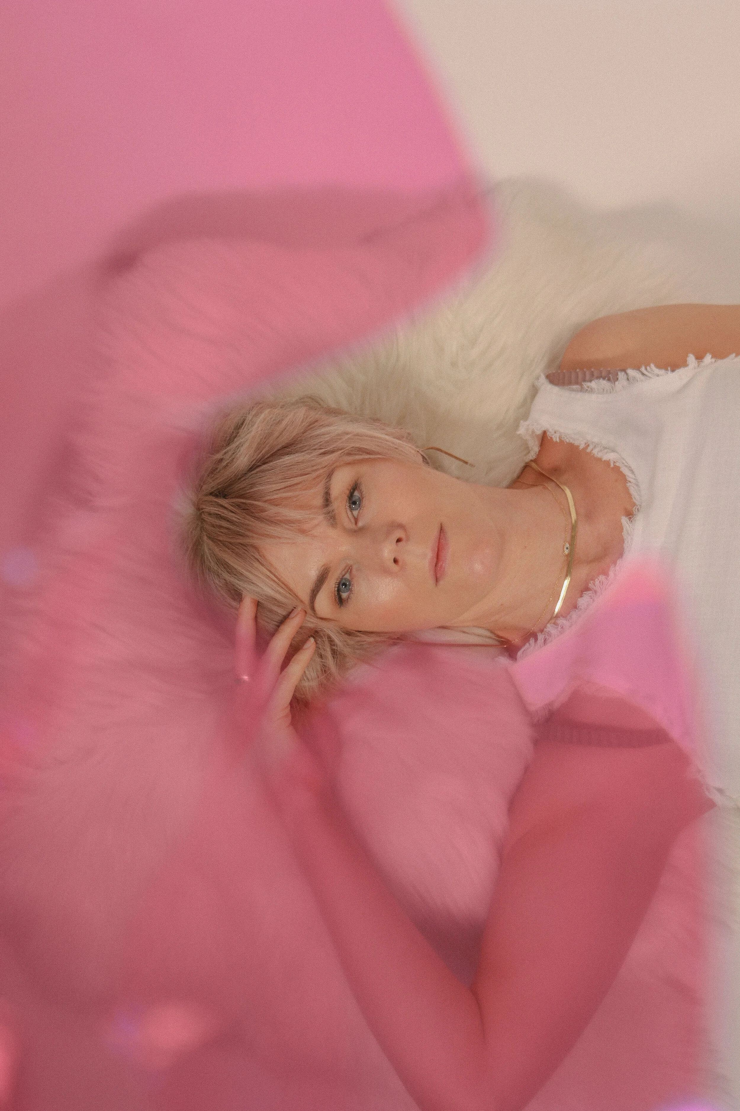 A young woman with blonde hair, blue eyes, and light skin lies on pink fluffy fabric, wearing a white distressed t-shirt and gold jewelry.