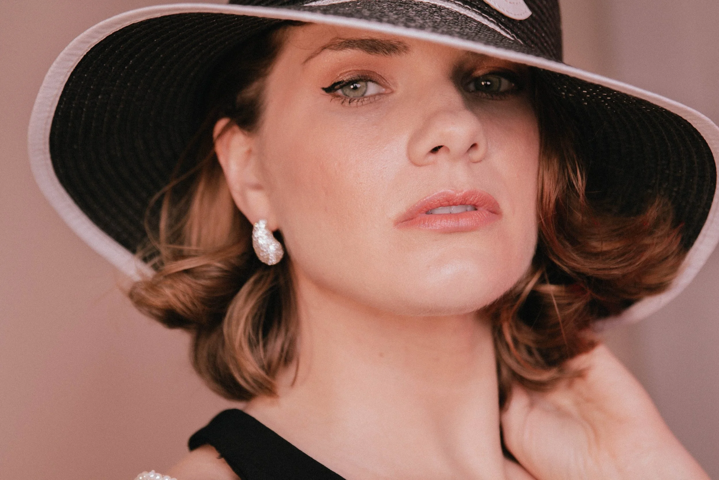 A woman wearing a black and white wide-brimmed hat, pearl earrings, and a black top, with styled hair and subtle makeup, posing against a soft background.