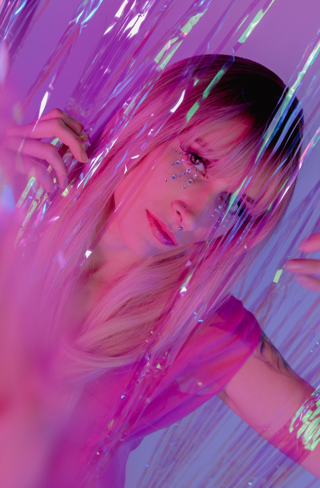 A close-up portrait of a woman with pink hair, artfully decorated with rhinestones around her eyes, partially obscured by iridescent, colorful paper strips.