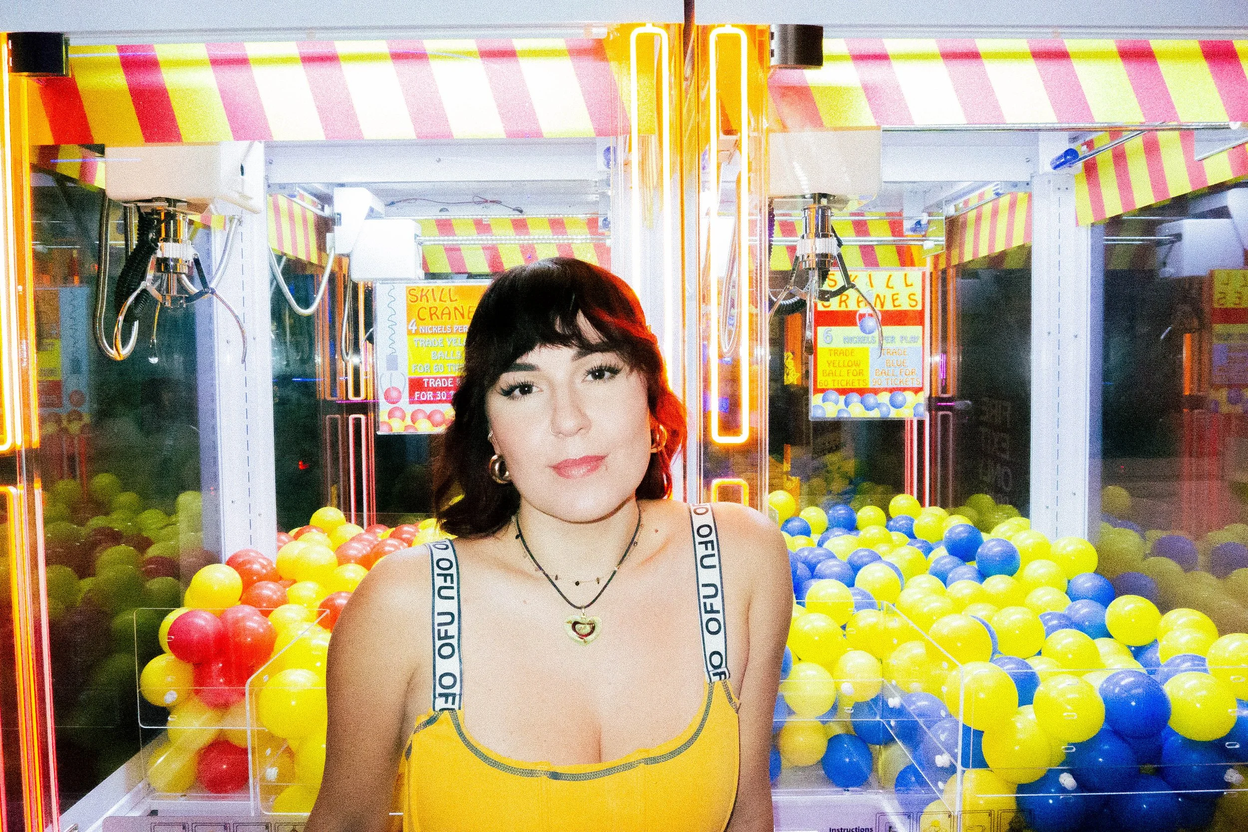 A woman with dark hair and earrings standing in front of a ball pit at an arcade, with colorful yellow, blue, and red balls and signs about skill cranes in the background.