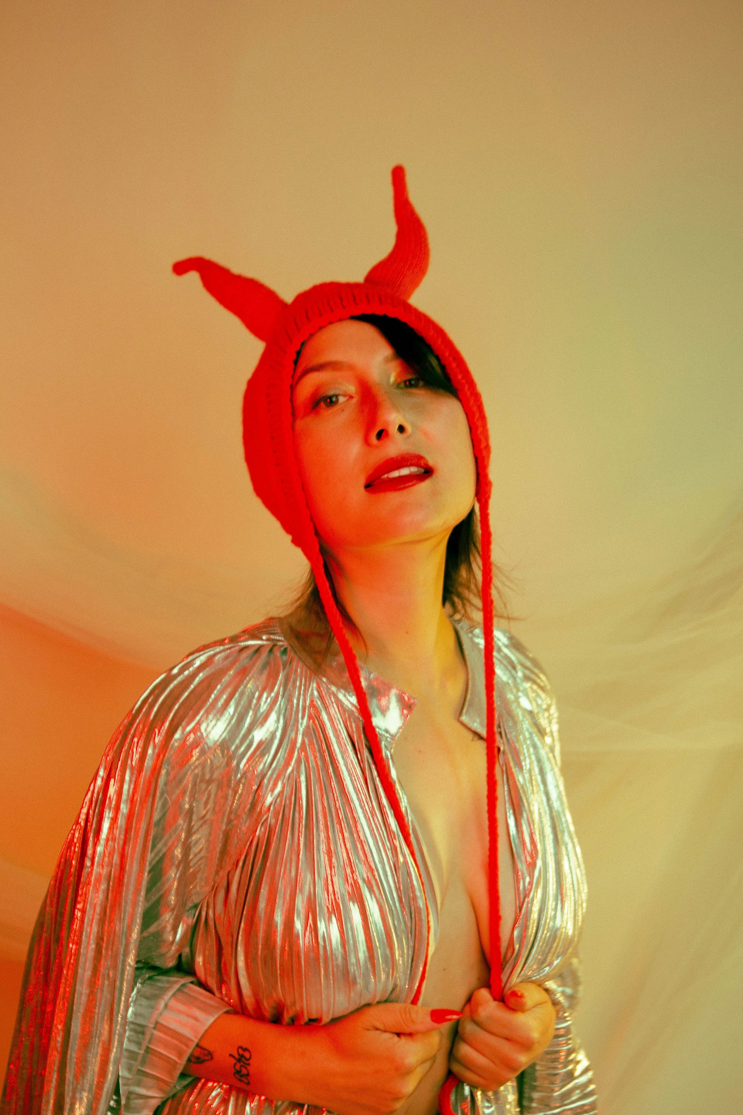 A person wearing a shiny, metallic, striped jacket and a red knit hat with horns. They have dark hair, fair skin, and red lipstick, and are standing against a plain, warm-toned background.