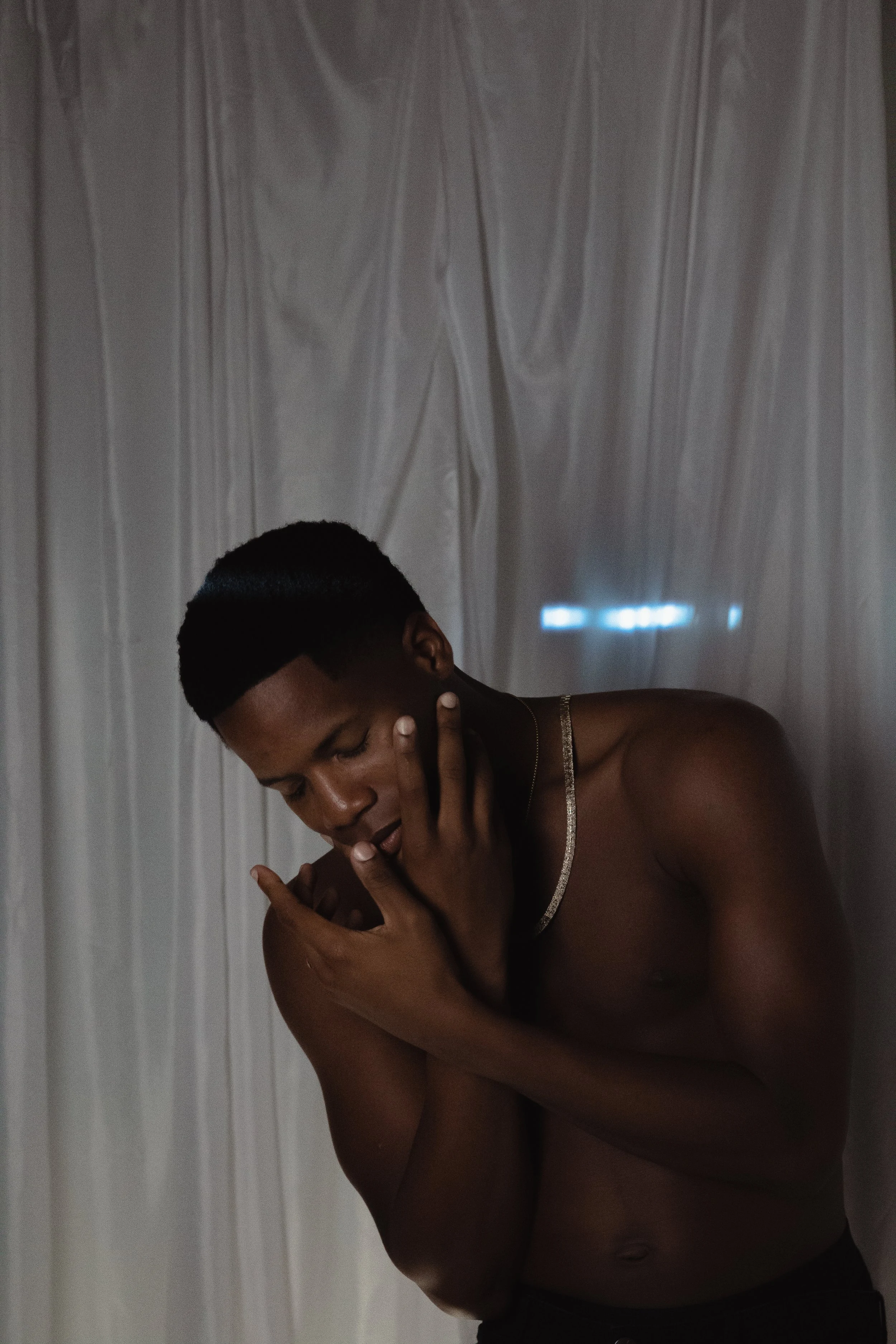 A shirtless man with a short hairstyle, standing against a plain curtain backdrop, covering part of his face with his hand, while his other hand rests on his chest.