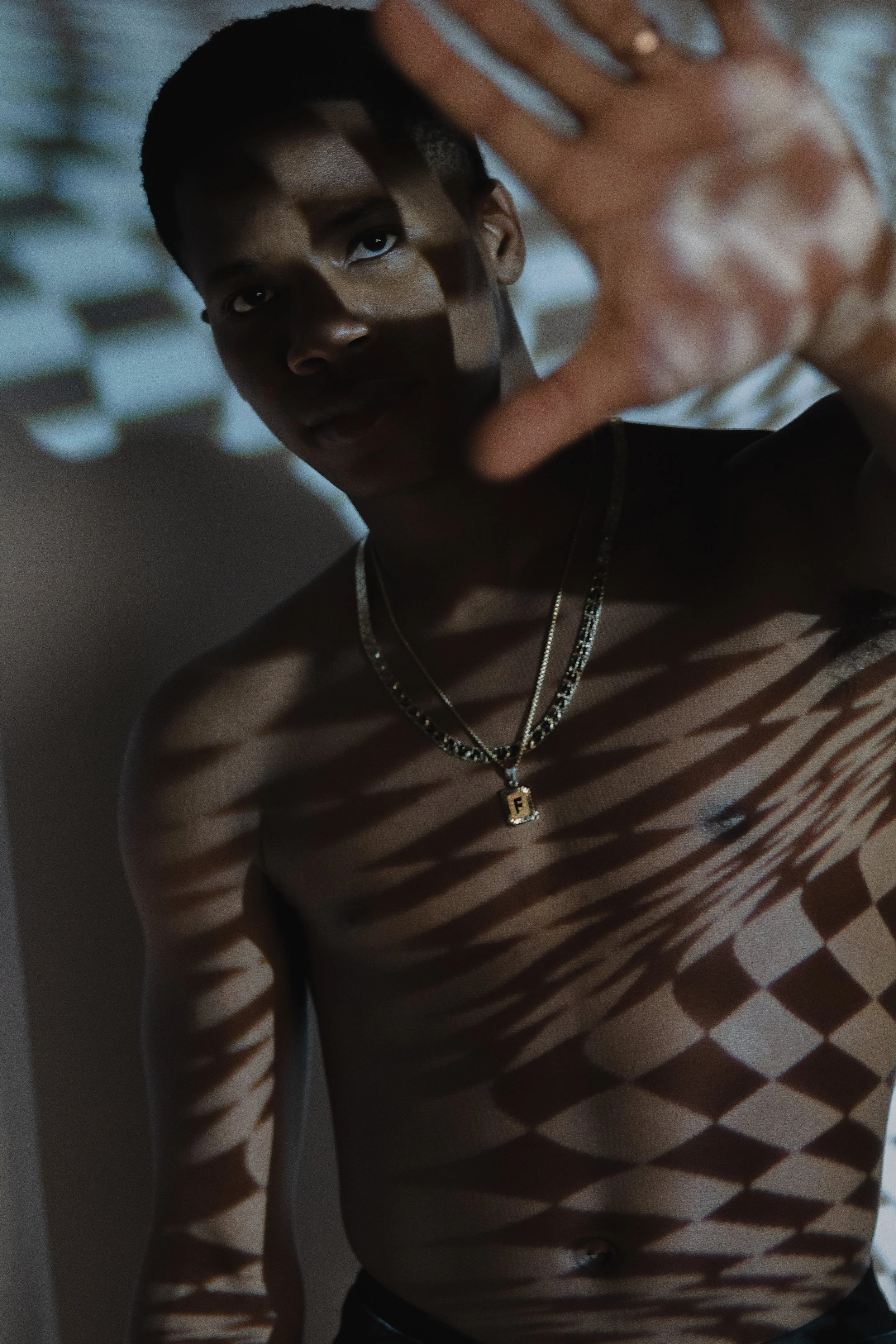 A person with dark skin and short hair, wearing layered necklaces including one with a pendant, standing indoors with striped and diamond-shaped light patterns cast on their body from a window blind. They are raising one hand toward the camera.