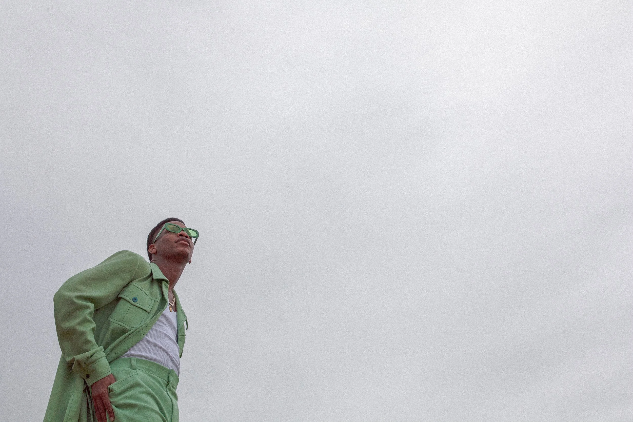 Young man in light green jacket and matching pants, wearing green sunglasses, standing outdoors against a cloudy gray sky.