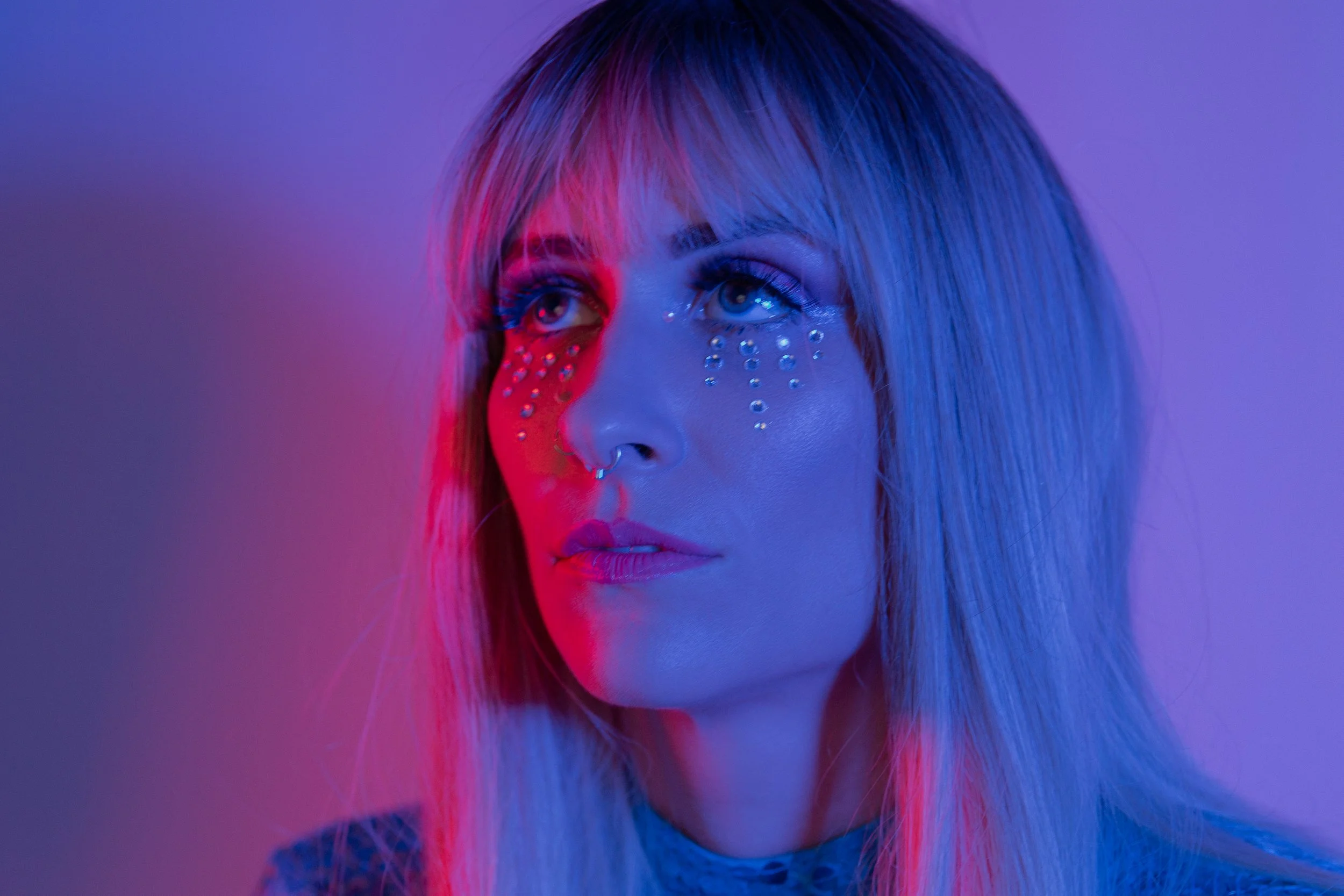 Close-up of a woman with blonde hair, rhinestone facial decorations, and makeup, illuminated with pink and blue lighting.